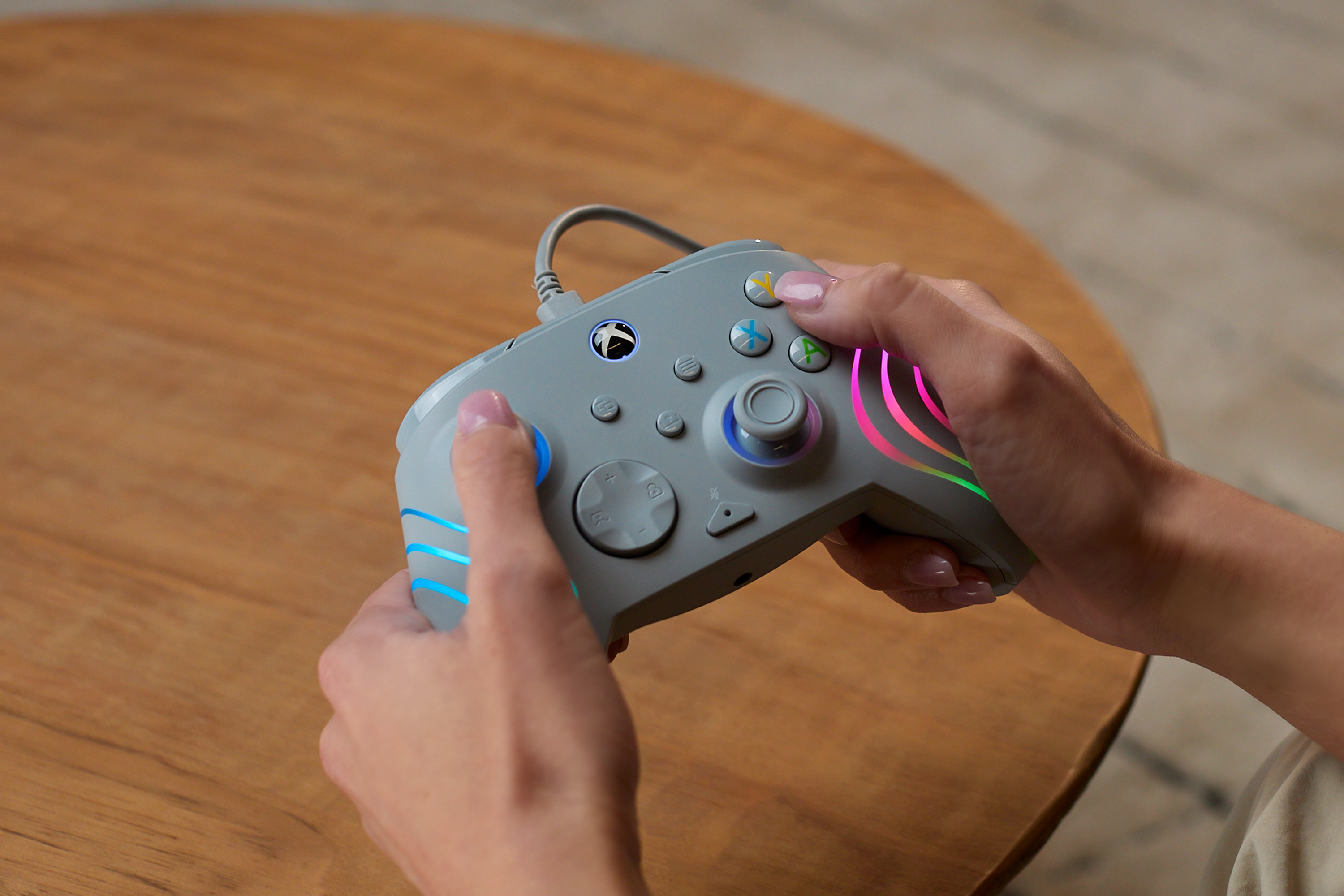 Personne tenant une manette Xbox grise avec des bandes LED colorées sur une table en bois.
