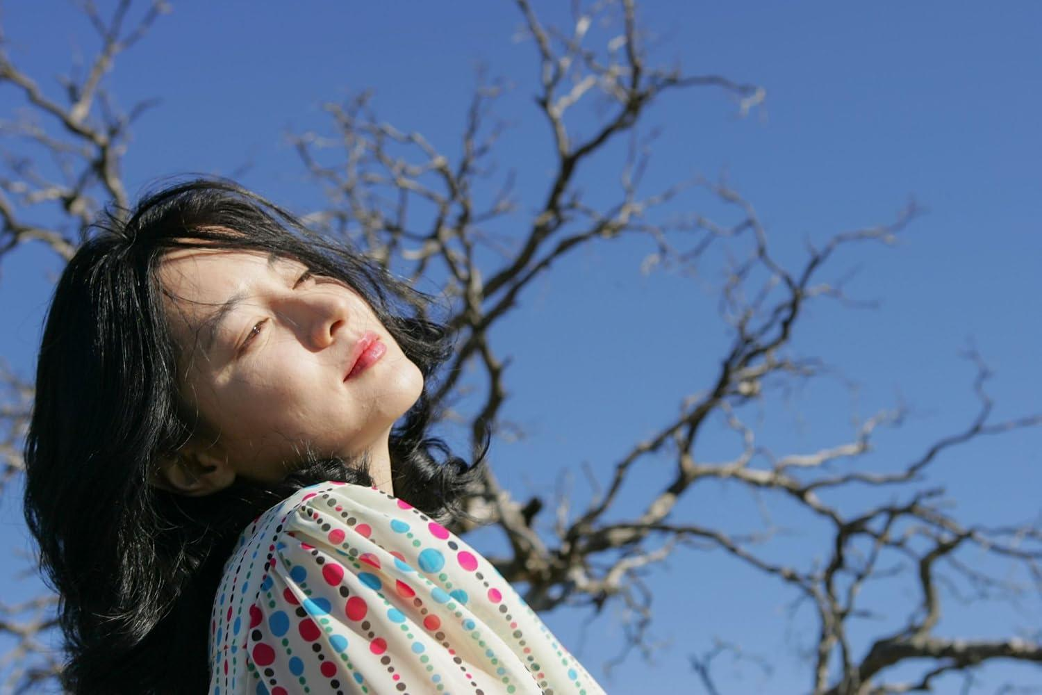 Eine Frau mit schwarzen Haaren und buntem Oberteil blickt in die Sonne, im Hintergrund kahle Äste und blauer Himmel.