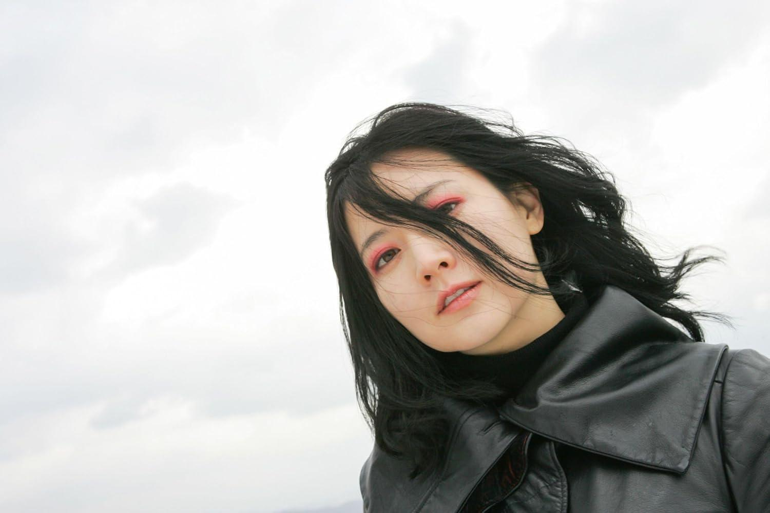 Eine Frau mit schwarzem Haar und rotem Lidschatten trägt eine schwarze Lederjacke vor einem grauen Himmel.