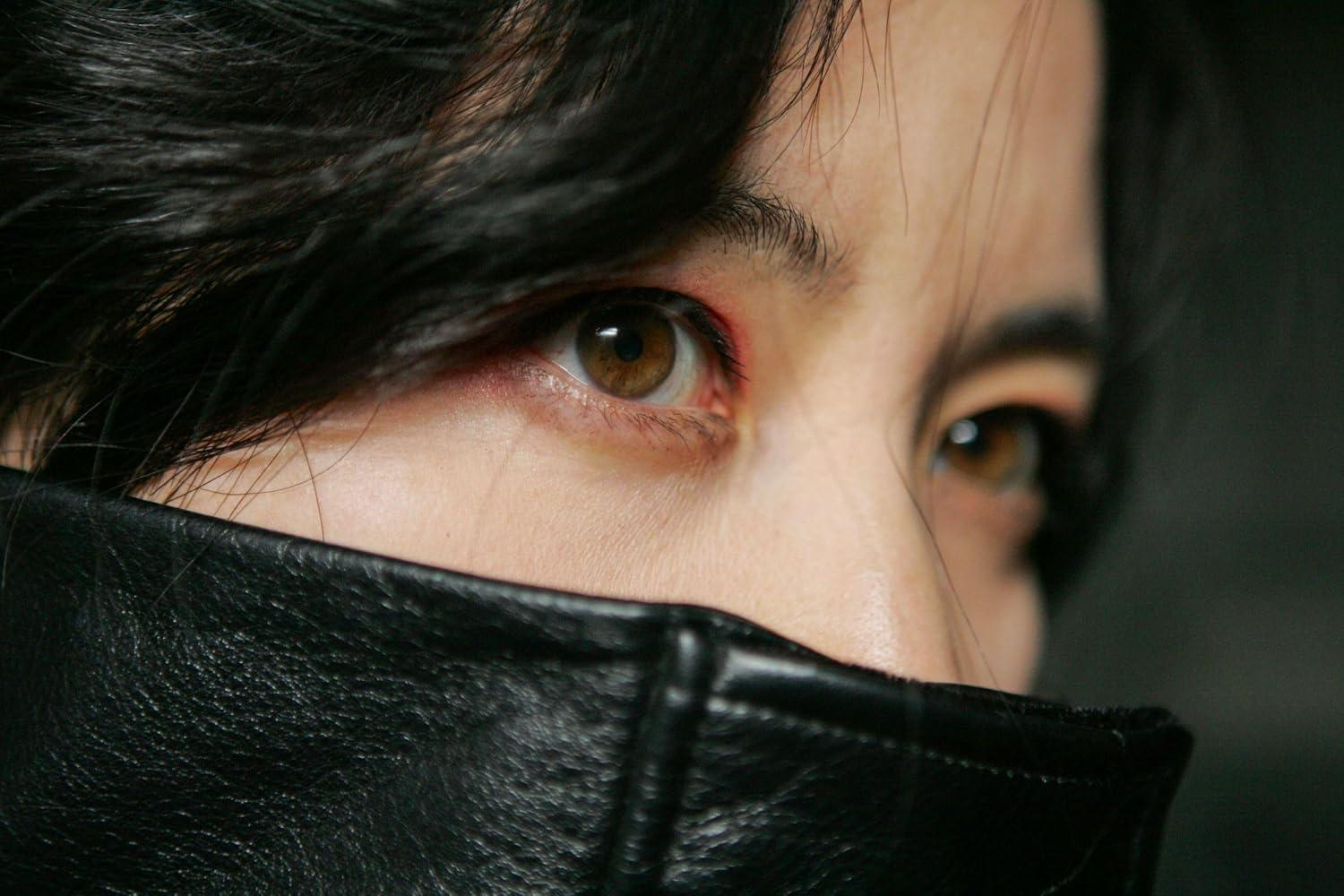 Nahaufnahme einer Person mit dunklem Haar, braunen Augen und einem schwarzen Lederkragen, der das untere Gesicht verdeckt.