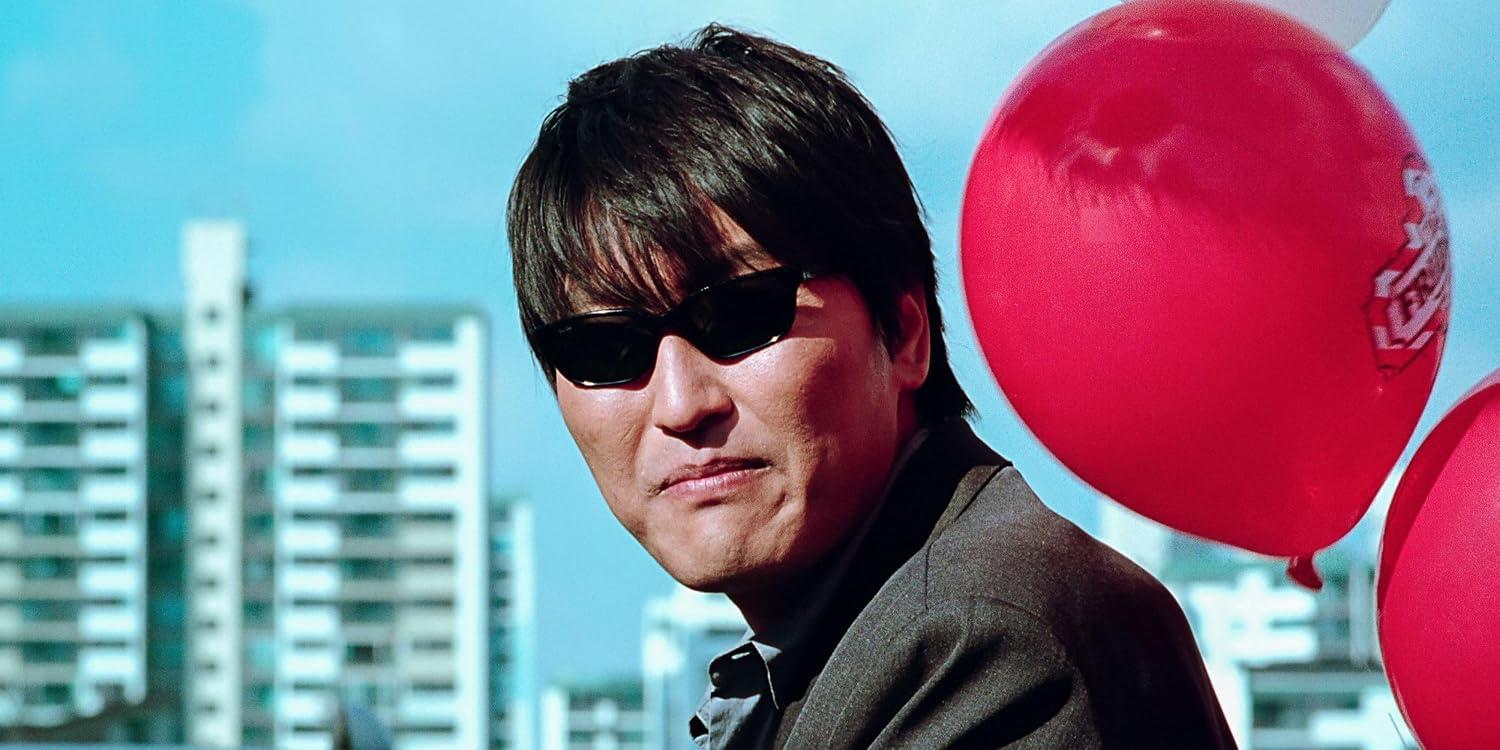 Ein Mann mit Sonnenbrille und Anzug posiert im Freien mit roten Ballons im Hintergrund.