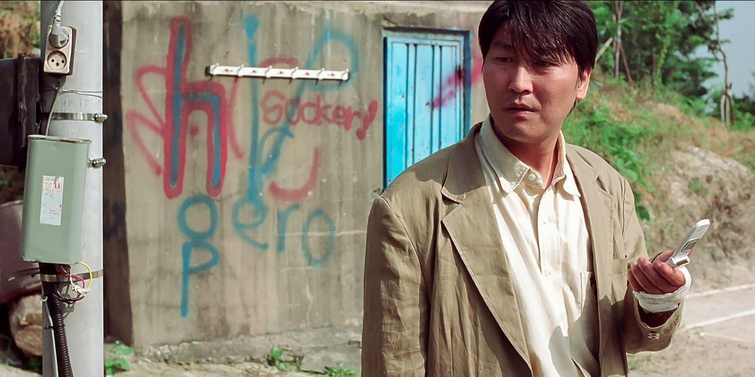 Ein Mann in einer beige Jacke mit Handy vor einer mit Graffiti bedeckten Wand.