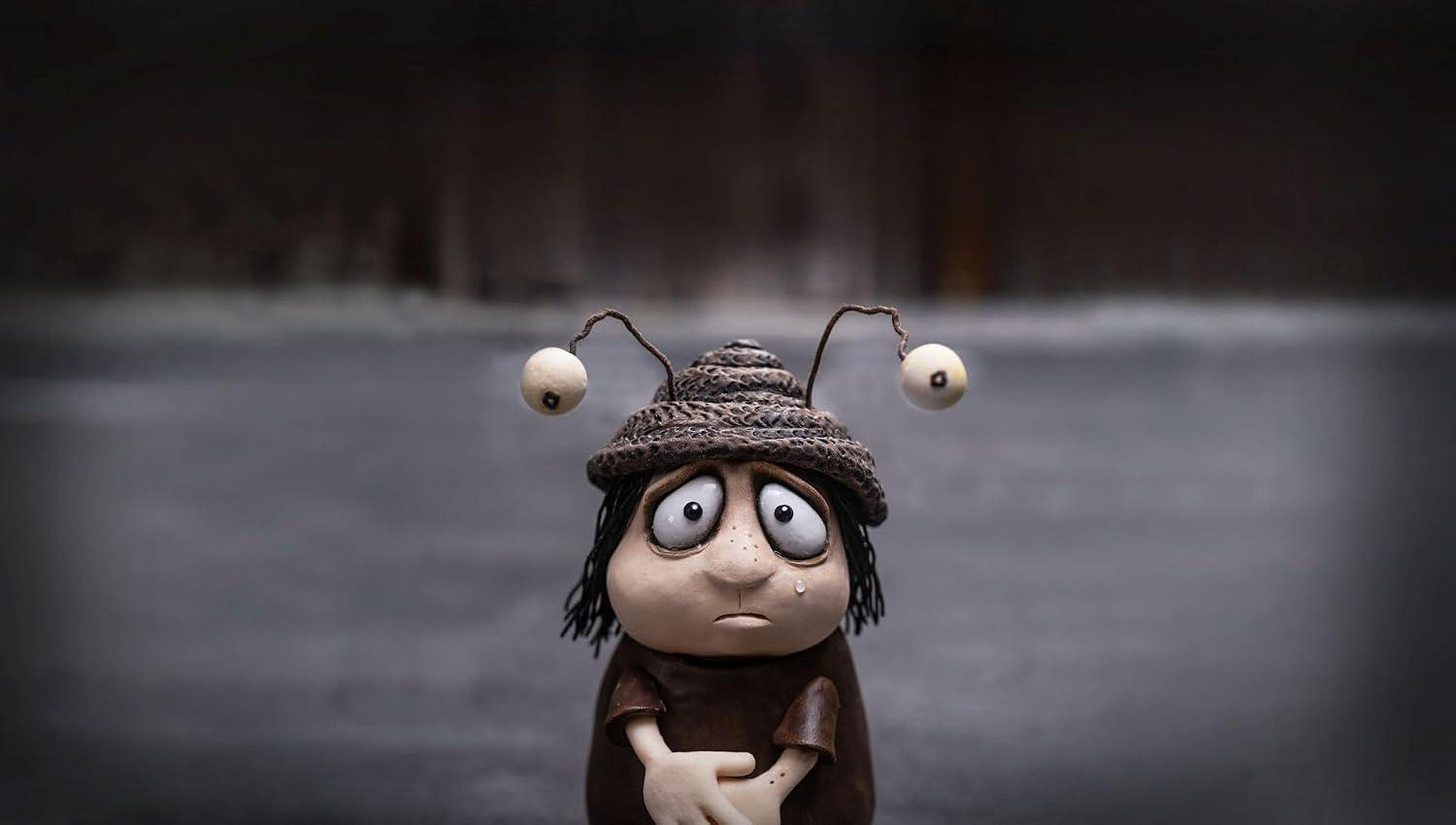 Eine traurige Tonfigur mit insektenartigen Augen auf Stielen, einem Strickhut und einer Träne auf dunklem Grund.