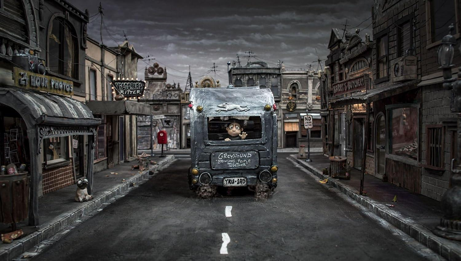 Ein Spielzeugbus fährt durch eine düstere Stadtstraße mit Geschäften unter einem bewölkten Himmel.