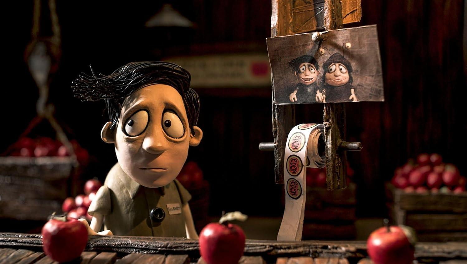 Stopp-Motion-Puppenfigur mit großen Augen an einem Apfelstand mit Schildern und roten Äpfeln im Hintergrund.