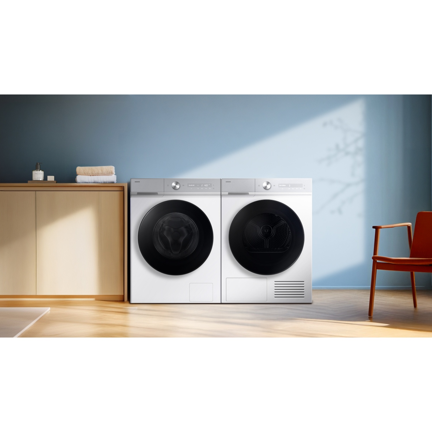 A white washing machine and dryer side-by-side in a light blue room with wooden cabinets and a red chair.