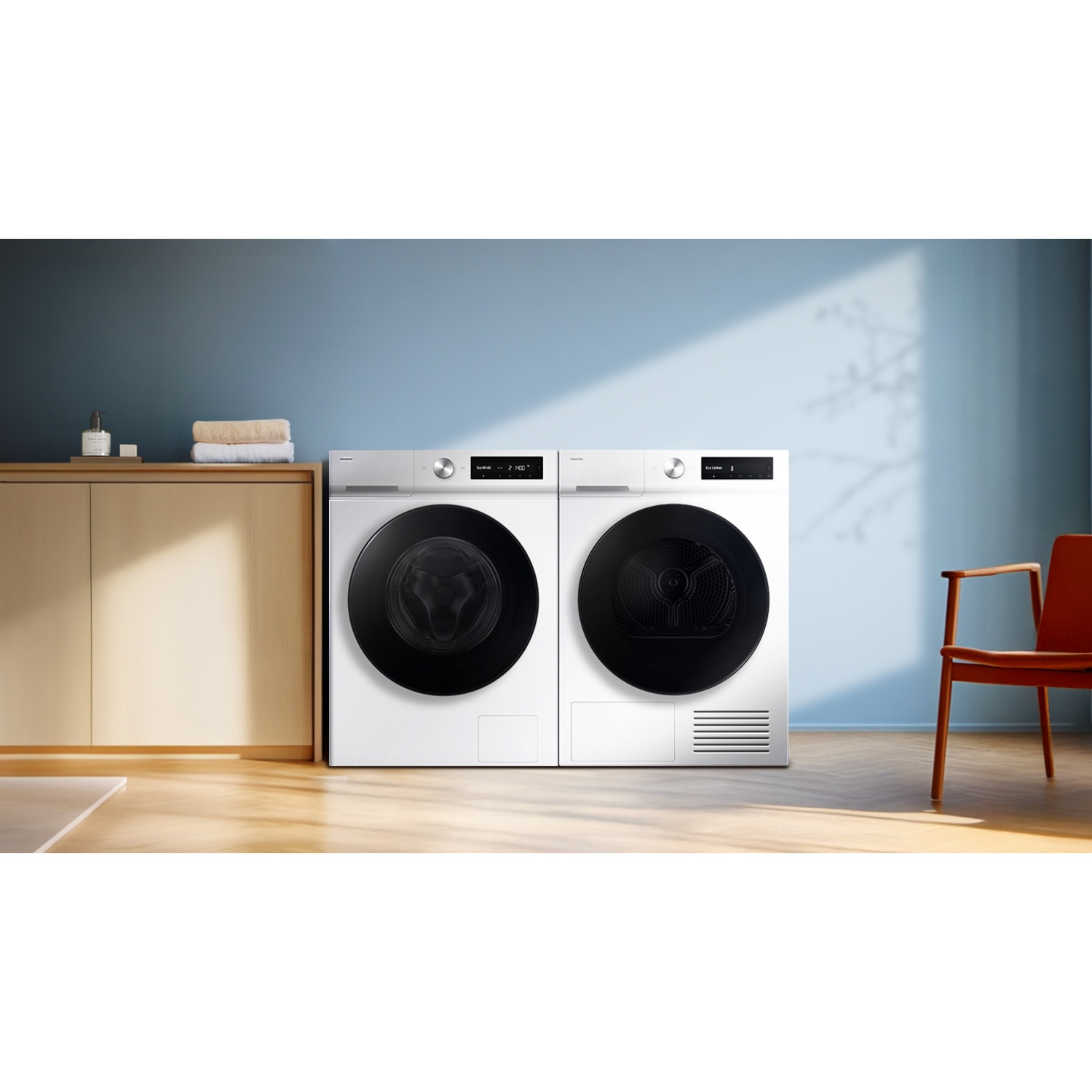 Witte wasmachine en droger naast elkaar in een moderne kamer met houten vloeren, een houten kast en een oranje stoel.
