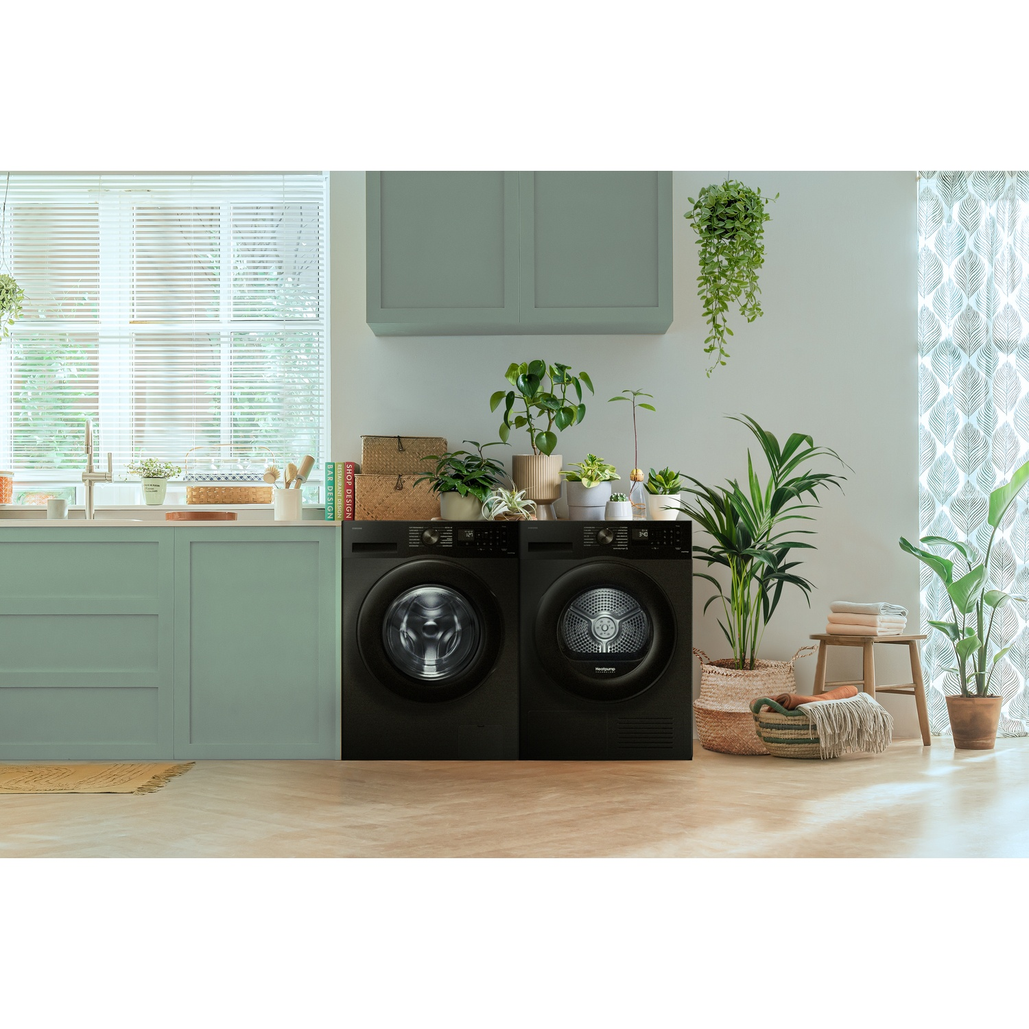 Two black laundry appliances stand in a light green room filled with various potted plants.