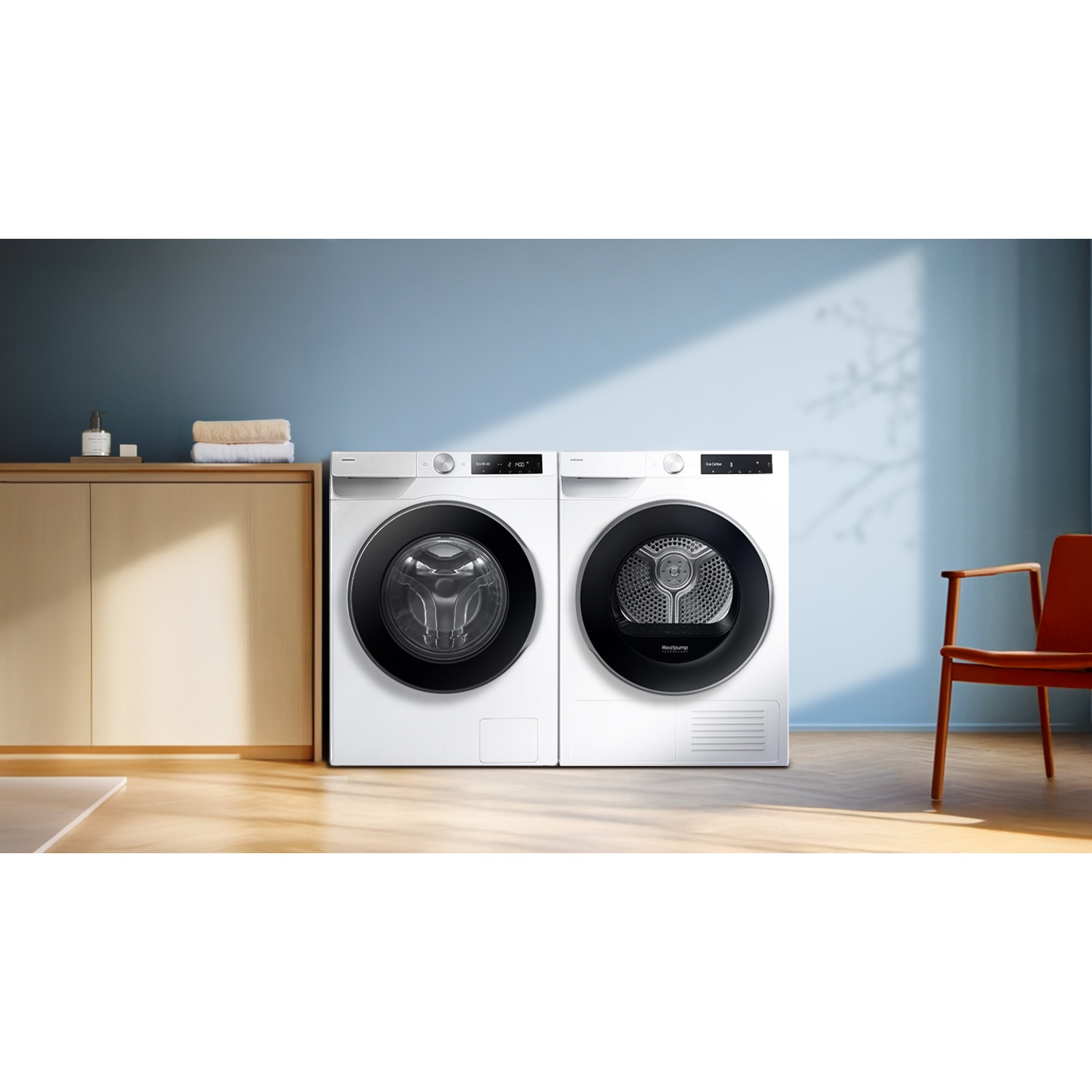 Witte wasmachine en droger naast elkaar in een kamer met blauwe muren, houten kast en een rode stoel.