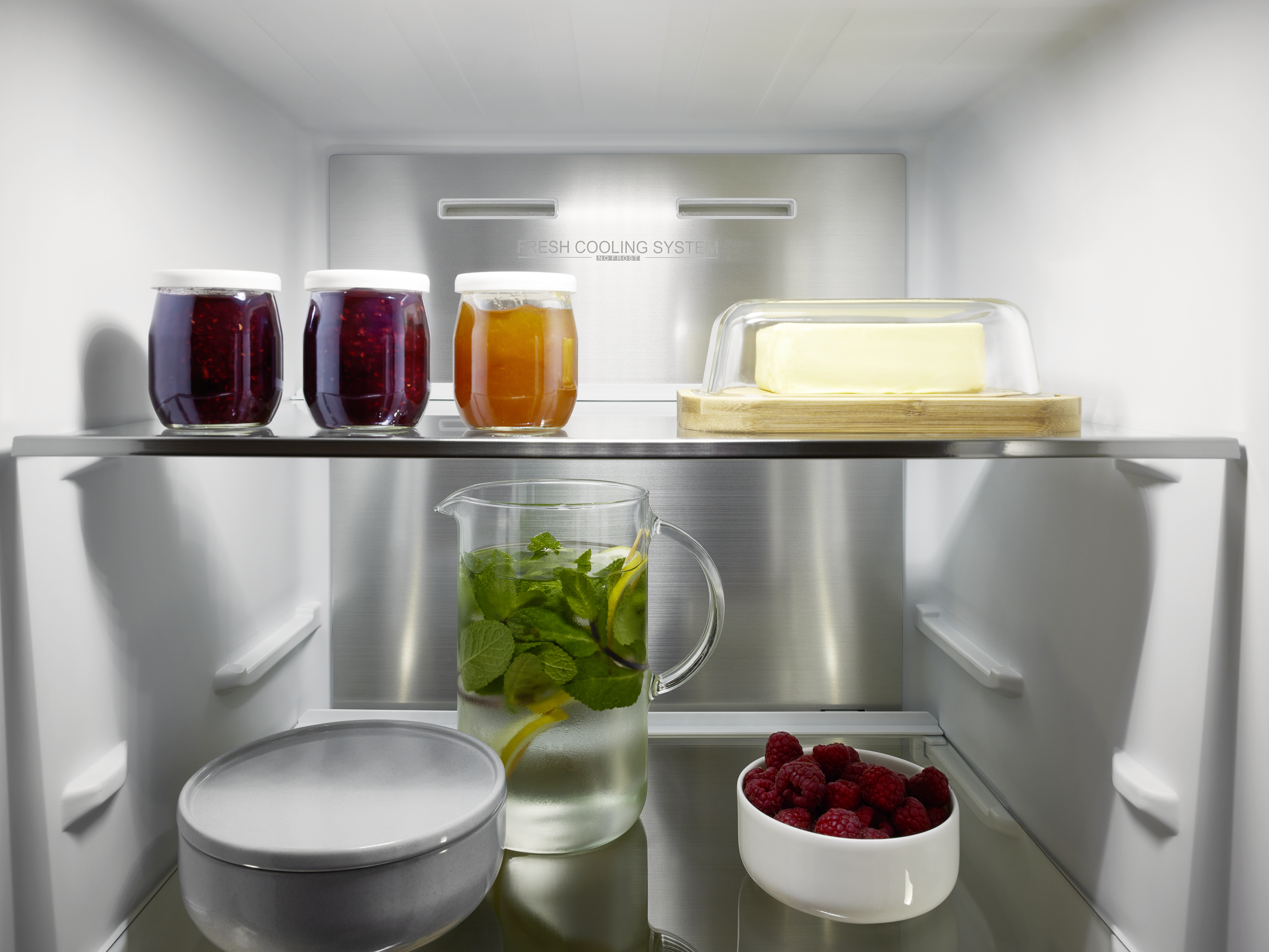 Ein ordentlicher Kühlschrank mit Gläsern voller Marmelade, Butter, einem Krug mit Minzwasser und einer Schale Himbeeren.