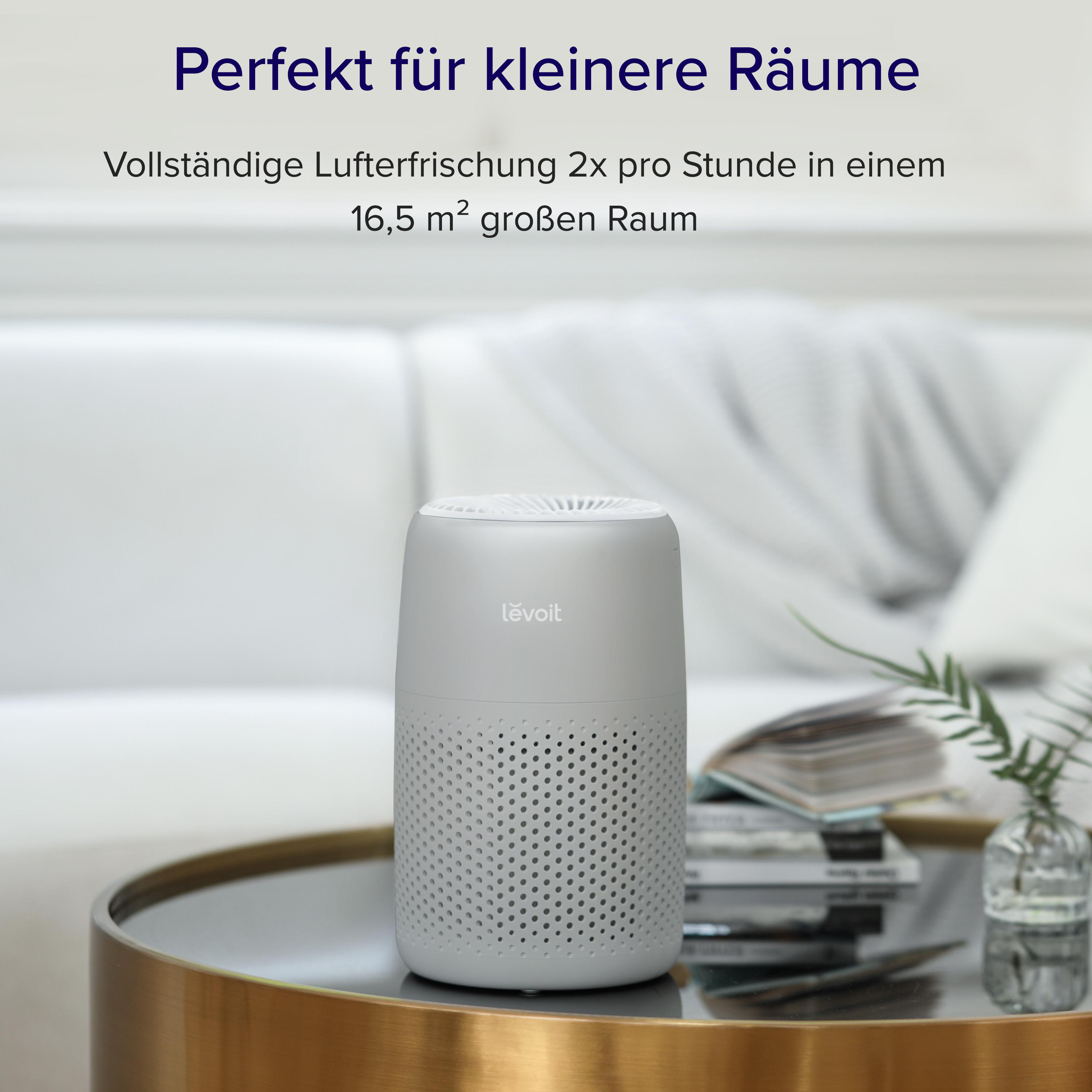 Grauer Levoit Luftreiniger auf einem runden Tisch in einem Wohnzimmer, mit Text oben.