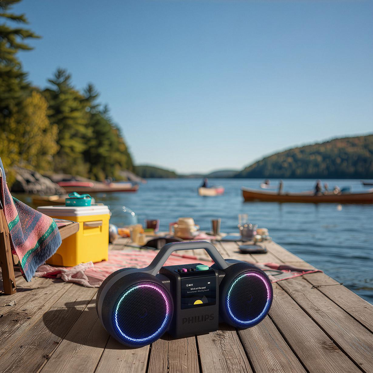Ein tragbarer Philips-Lautsprecher mit LED-Ringen steht auf einem hölzernen Steg am See mit Booten im Hintergrund.