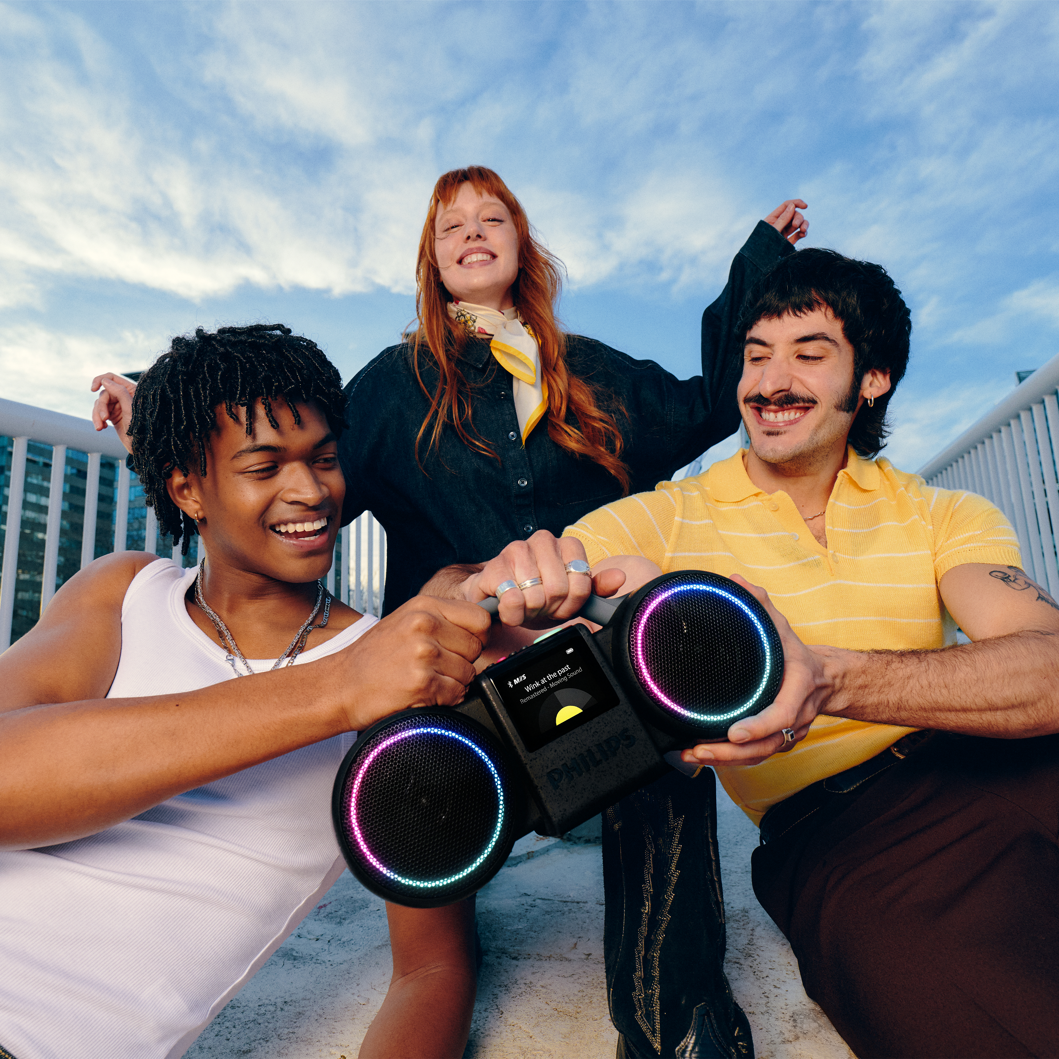 Drei lächelnde junge Erwachsene halten im Freien einen leuchtenden Philips Ghetto-Blaster.