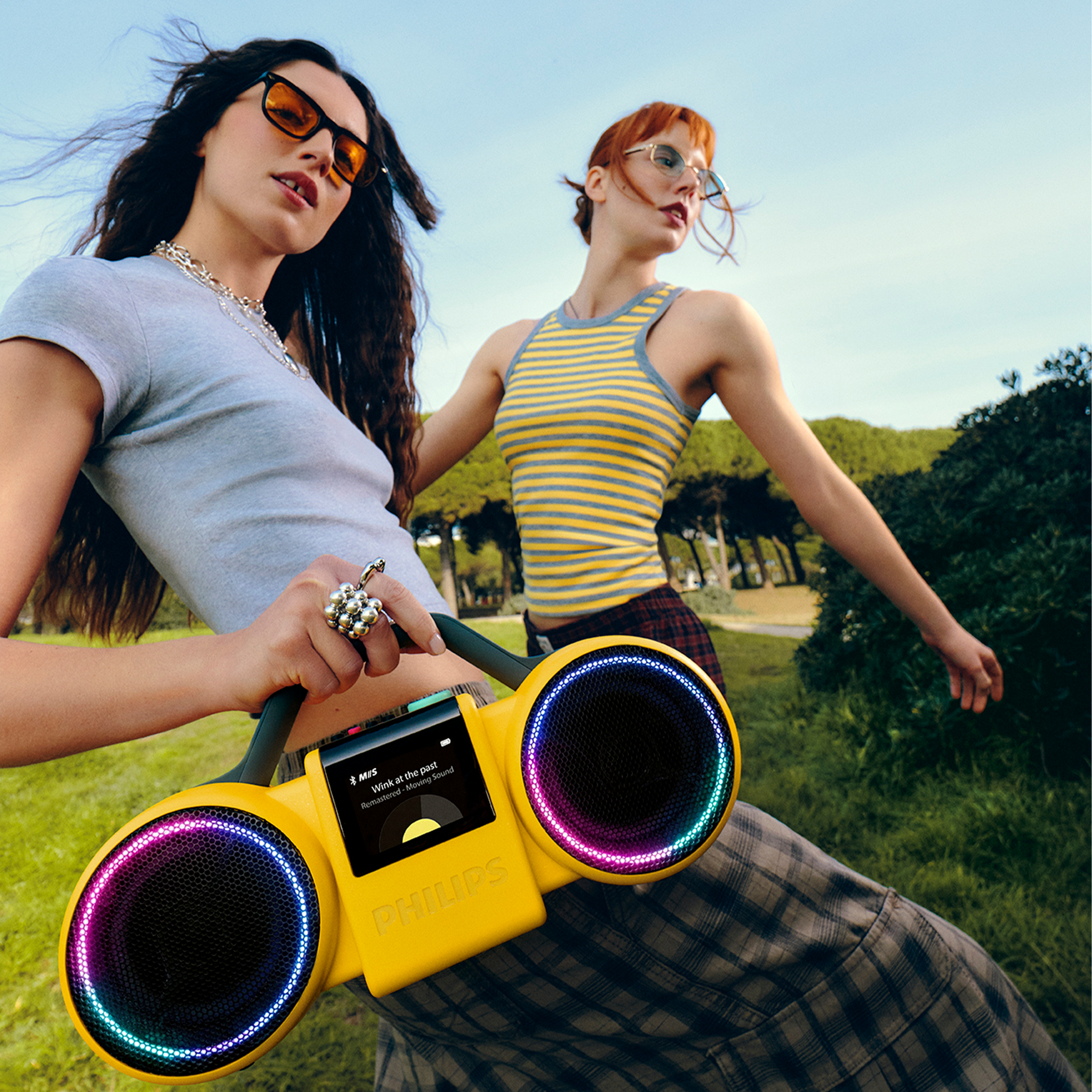 Zwei junge Frauen im Freien mit einem gelben Philips Boombox-Lautsprecher mit buntem LED-Licht.