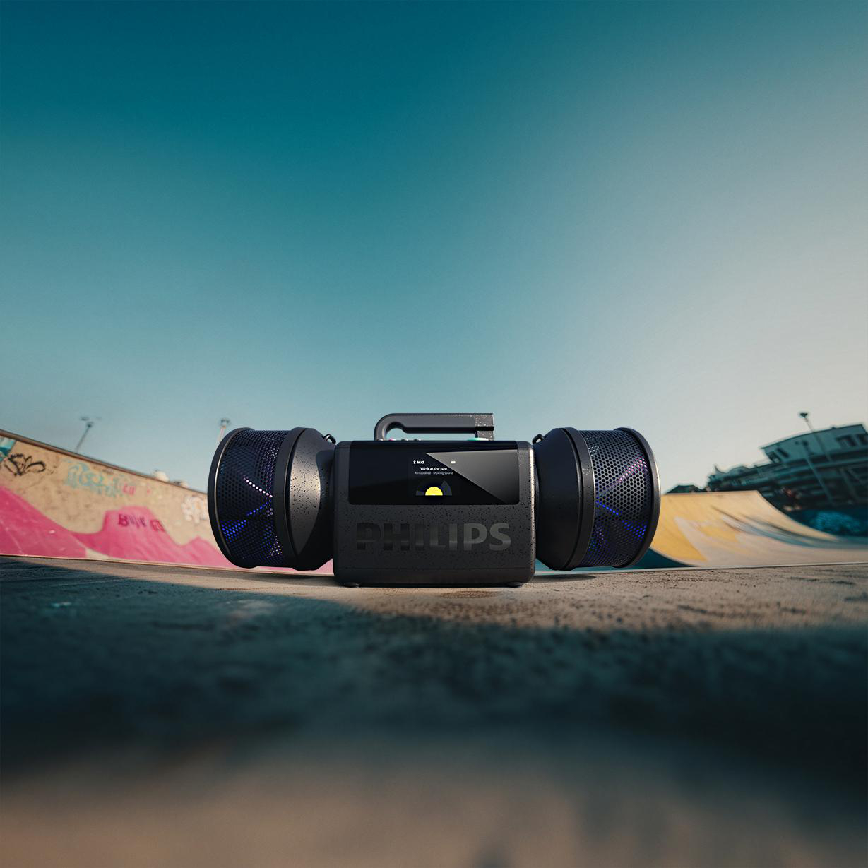 Ein schwarzer Philips Boombox-Lautsprecher steht im Freien auf Beton vor einer Skatepark-Rampe unter blauem Himmel.