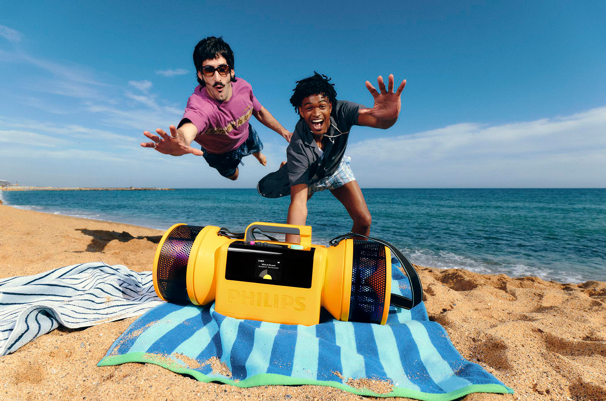 Zwei junge Männer springen fröhlich am Strand neben einem gelben Philips-Lautsprecher in die Luft.