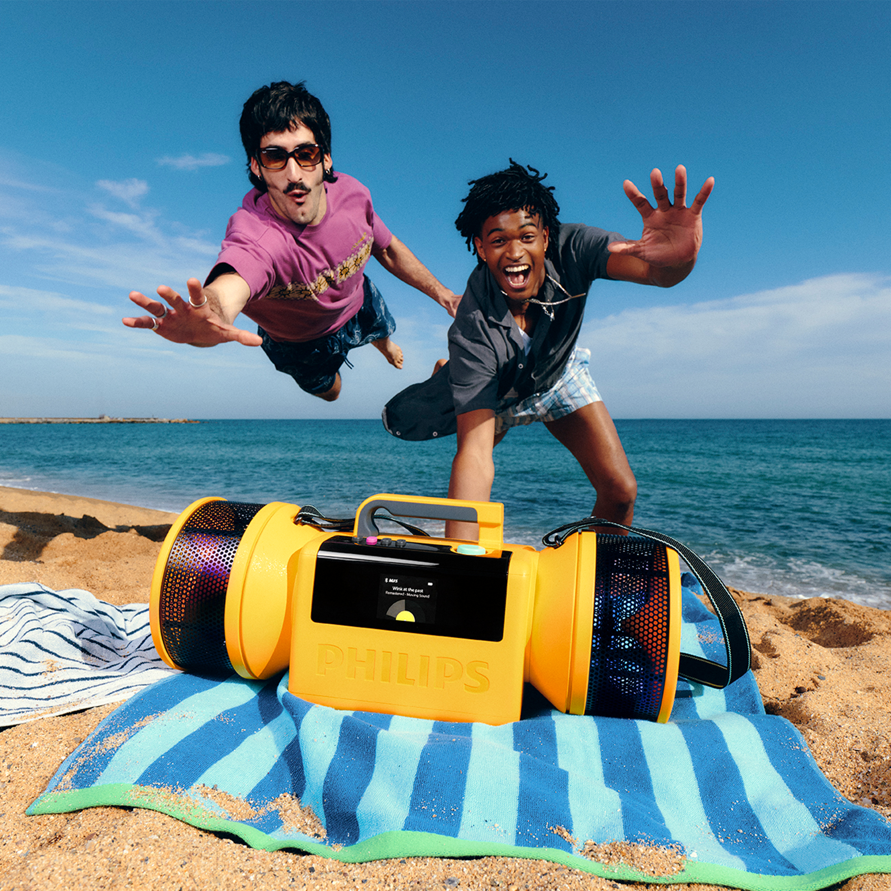 Zwei junge Männer jubeln ausgelassen am Strand hinter einem gelben Philips-Ghettoblaster auf einem blauen Handtuch.