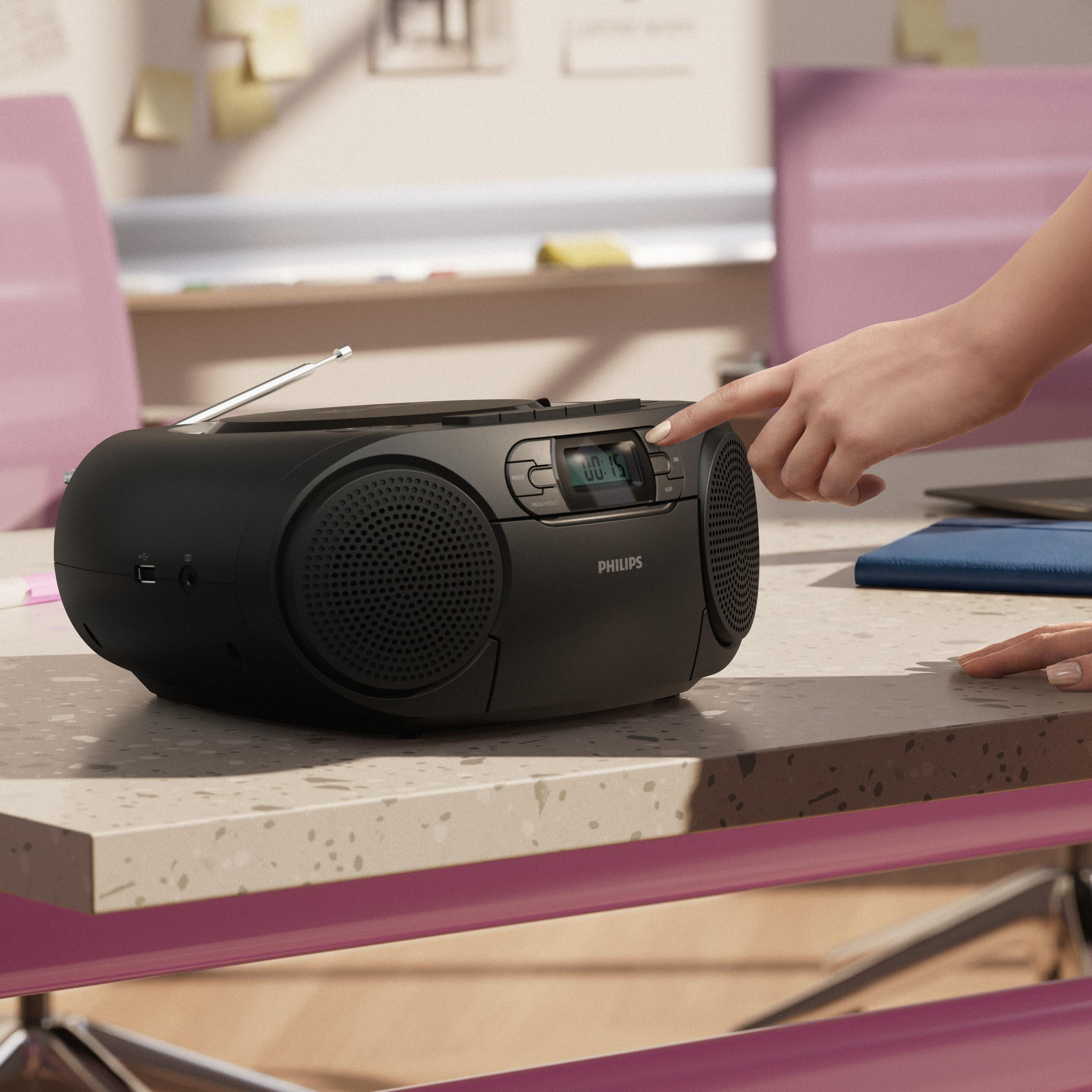 Eine Hand drückt die Taste eines schwarzen Philips-Radios, das auf einem Schreibtisch mit rosa Stühlen steht.
