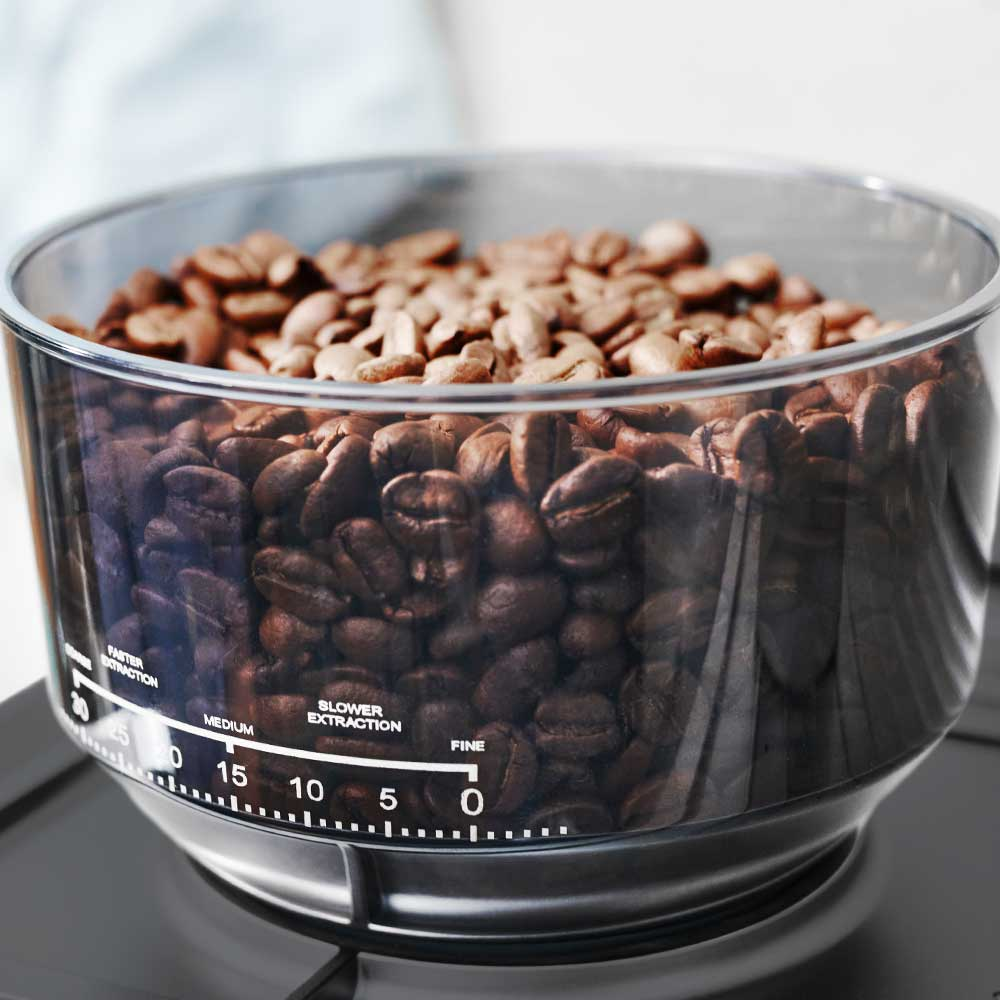 Transparenter Kaffeebohnenbehälter gefüllt mit braunen Kaffeebohnen, mit Mahlgradskala an der Vorderseite.