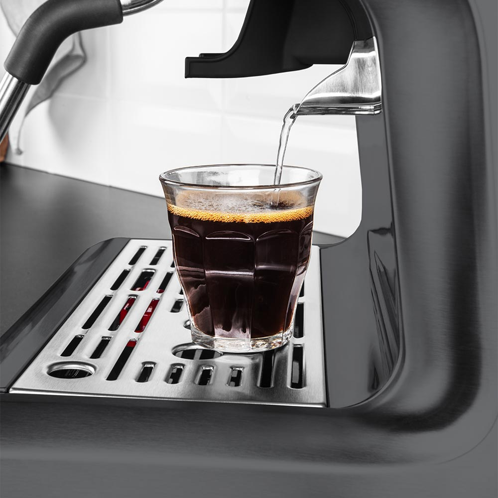 Eine Espressomaschine gießt heißes Wasser in ein kleines Glas auf einer silbernen Abtropffläche.
