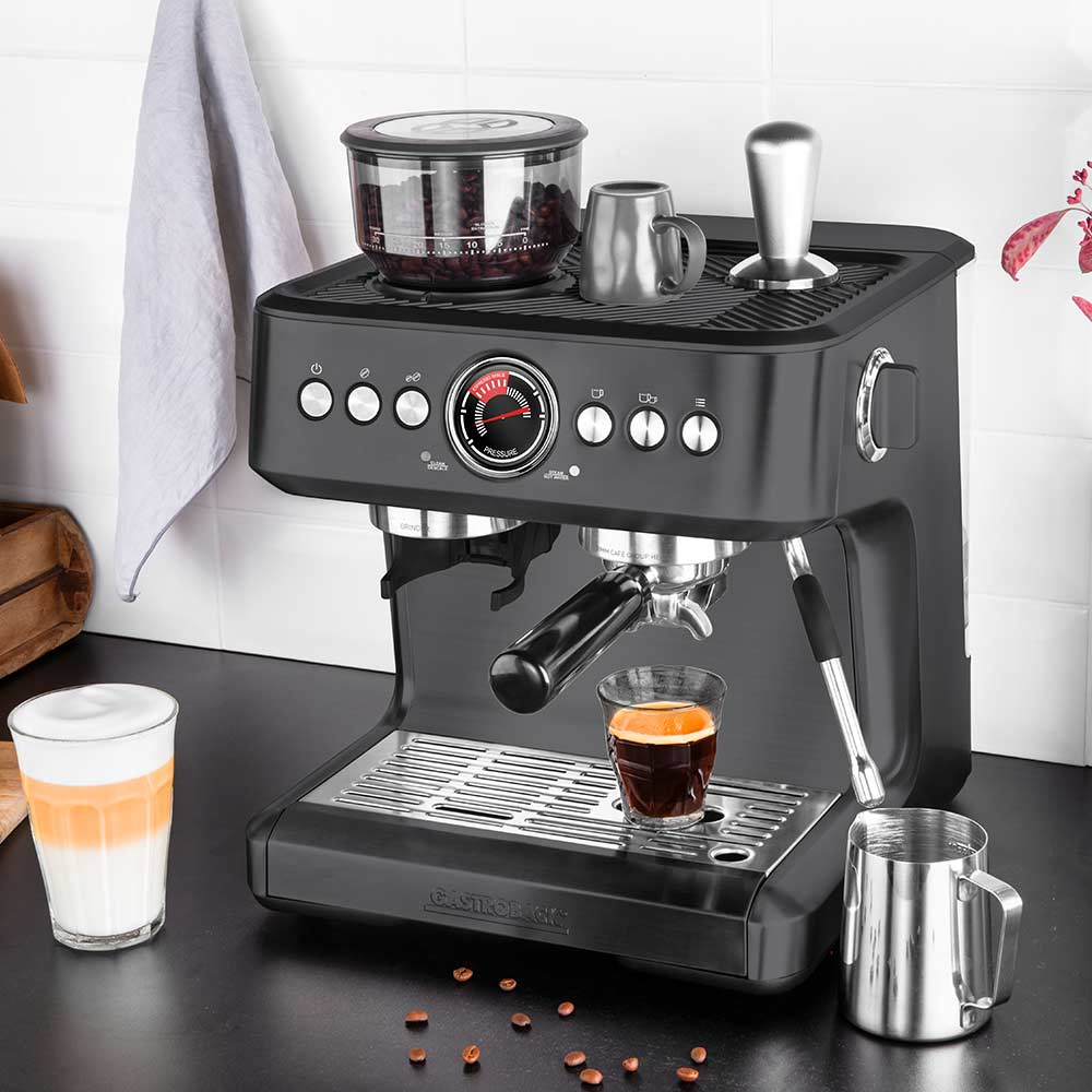 Eine schwarze Espressomaschine auf einer Theke mit Kaffeebohnen, einer Tasse Espresso, einem Milchglas und Zubehör.