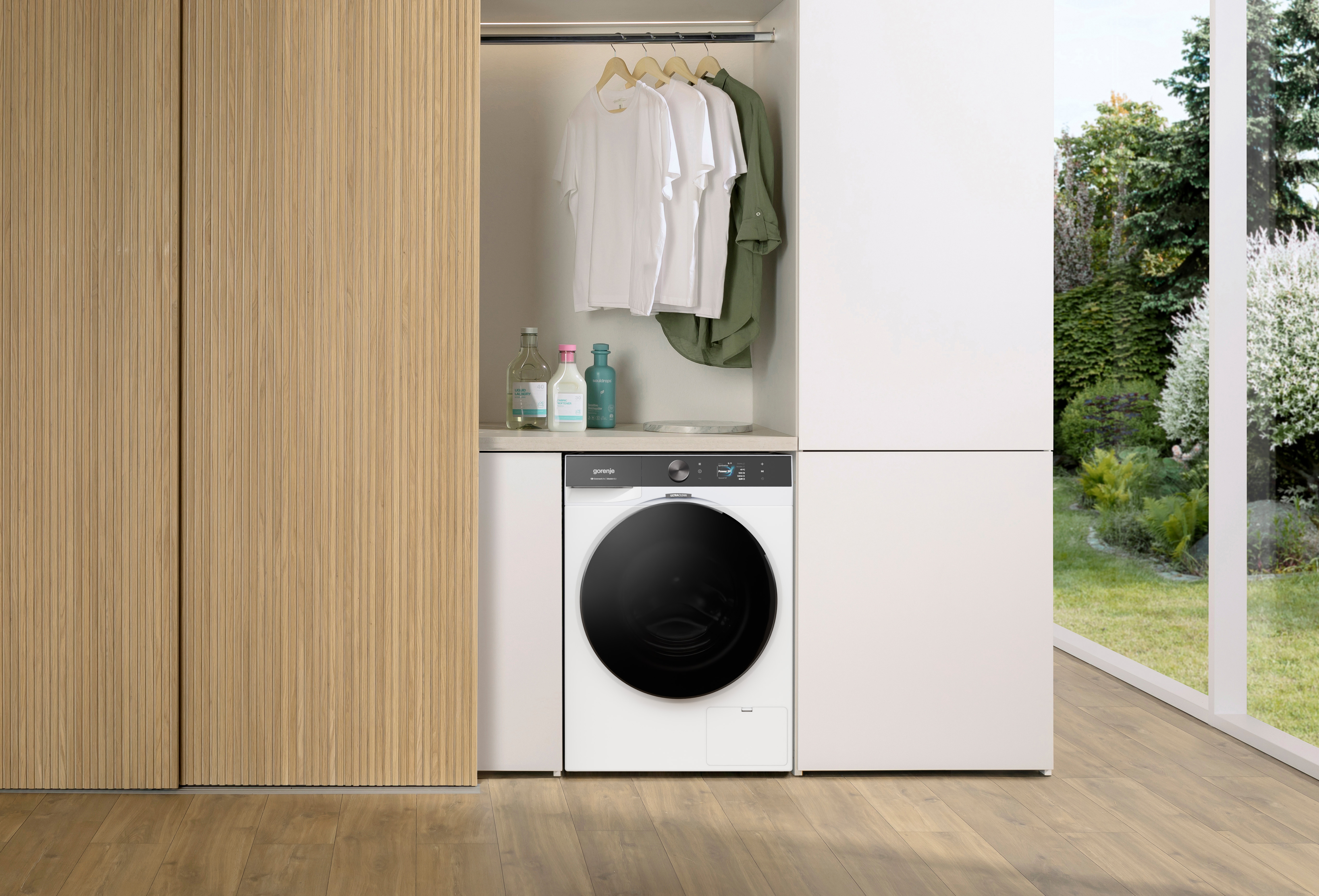 Eine weiße Waschmaschine in einer Holzschrankwand mit Hängekleidung und Reinigungsmitteln.