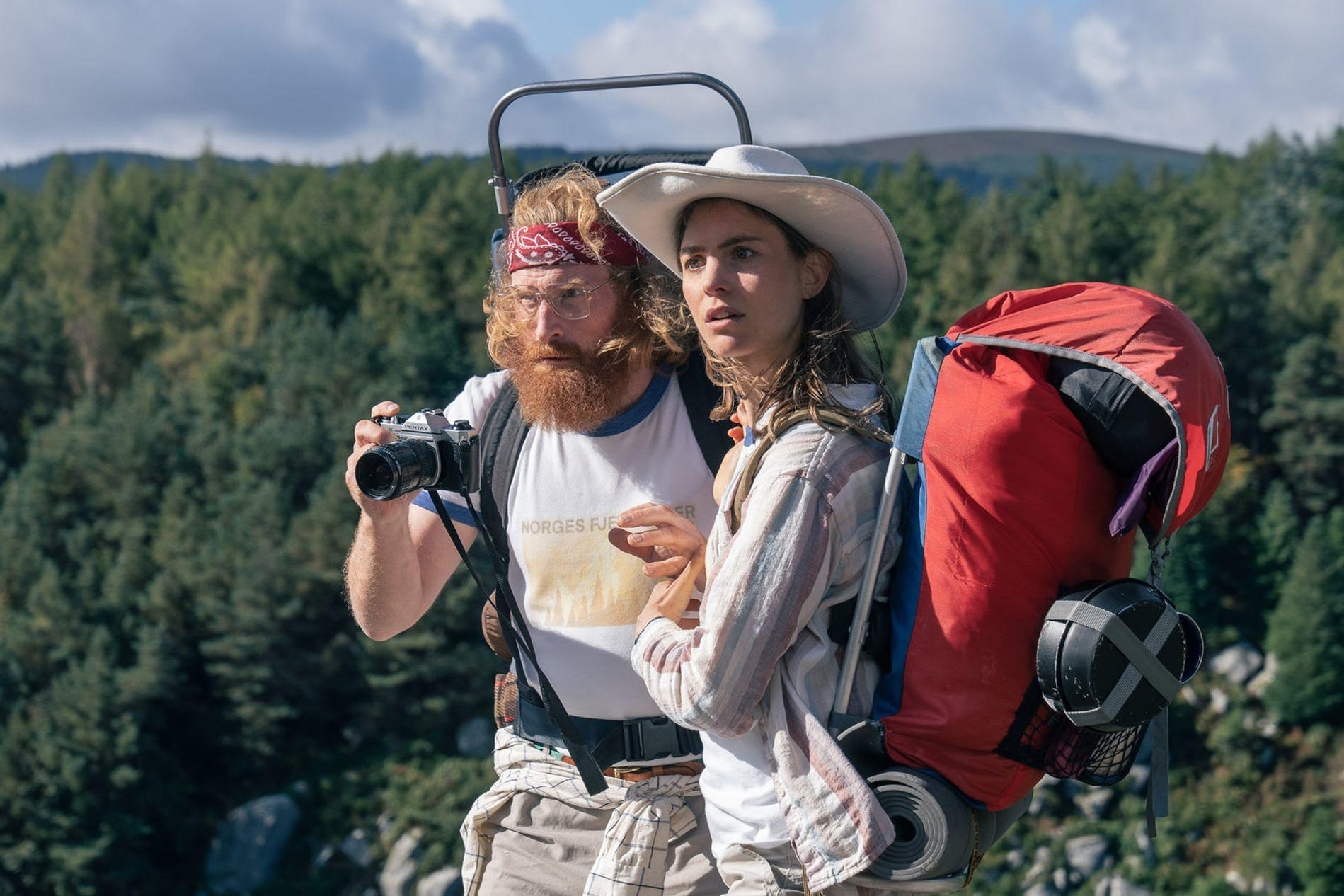 Zwei Wanderer mit großen Rucksäcken in einer waldreichen Berglandschaft; der Mann hält eine Kamera.