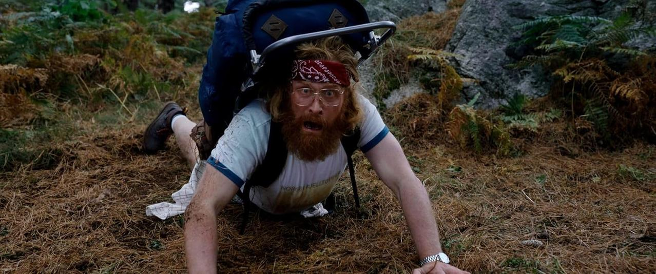 Ein Mann mit rotem Bart und Brille kriecht auf dem Waldboden und trägt einen großen Rucksack.