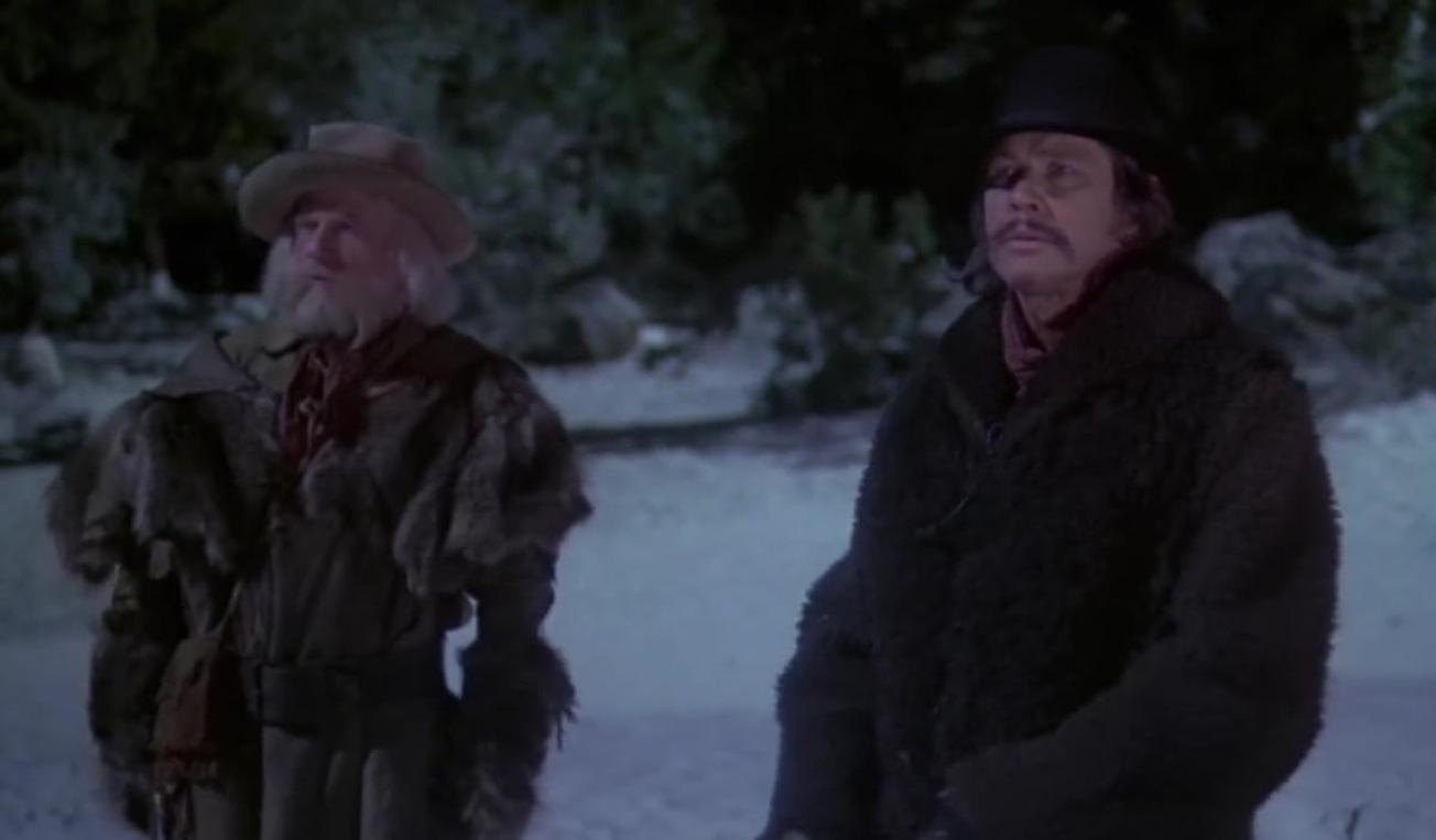 Zwei Männer in schweren Wintermänteln und Hüten stehen nachts draußen in einem schneebedeckten Wald.