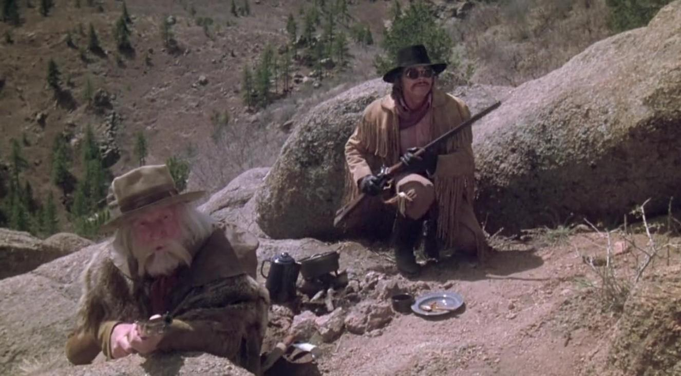 Zwei bewaffnete Männer im Western-Stil kauern in einer steinigen Berglandschaft mit Bäumen im Hintergrund.