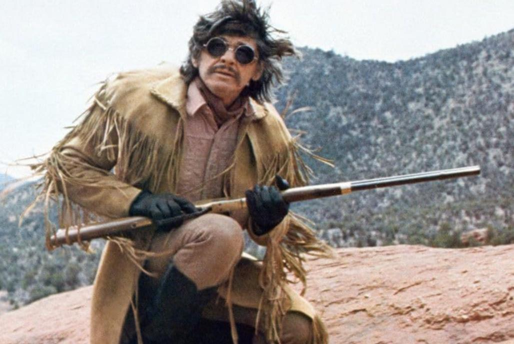 Ein Mann mit Fransenjacke, Sonnenbrille und Gewehr kniet in einer hügeligen Landschaft.