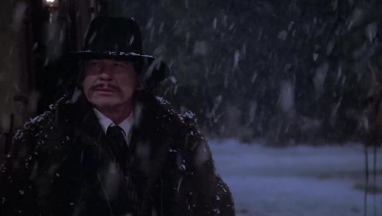 Ein Mann mit Hut und dunklem Mantel steht im starken Schneetreiben.