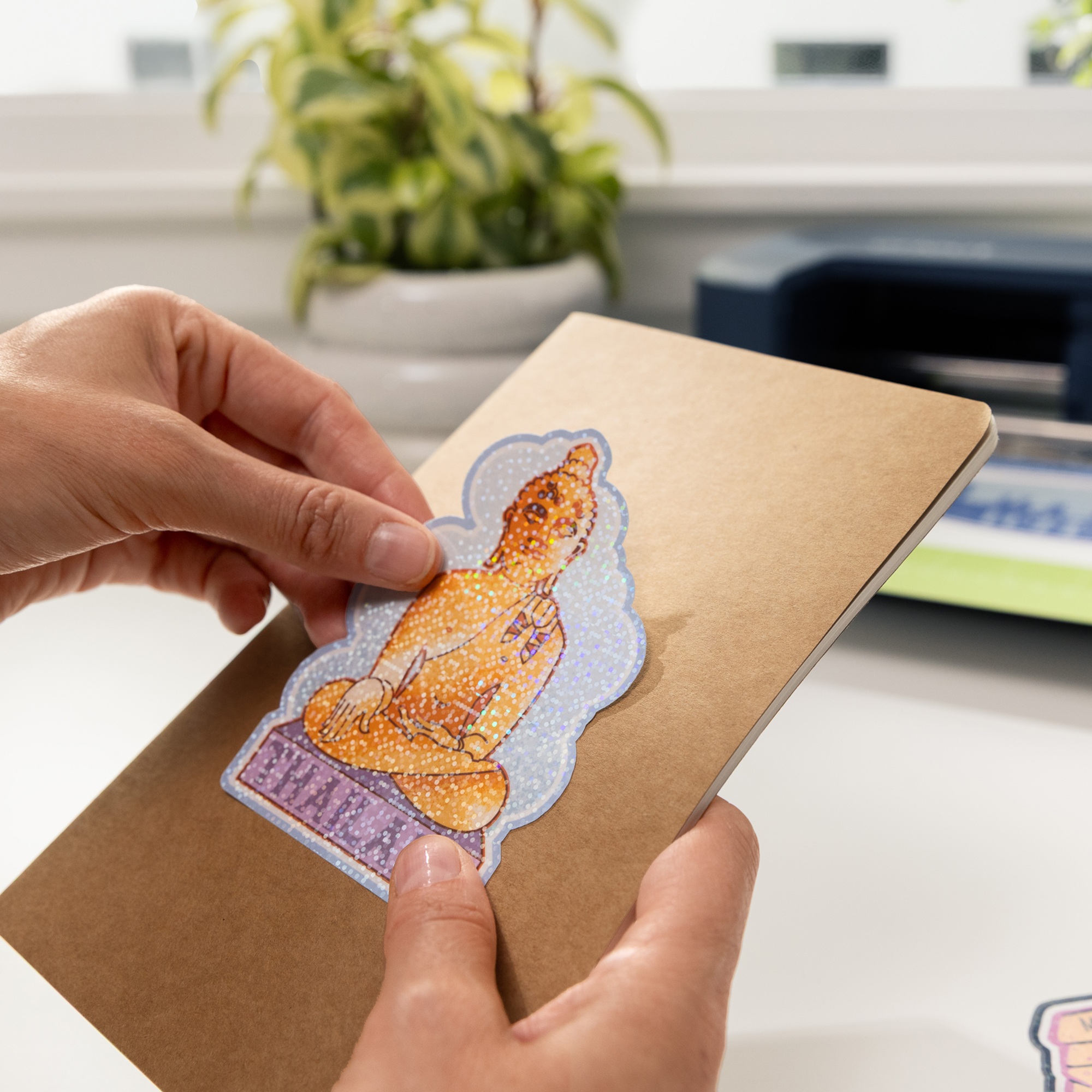 Una persona coloca una pegatina brillante de Buda en un cuaderno marrón.