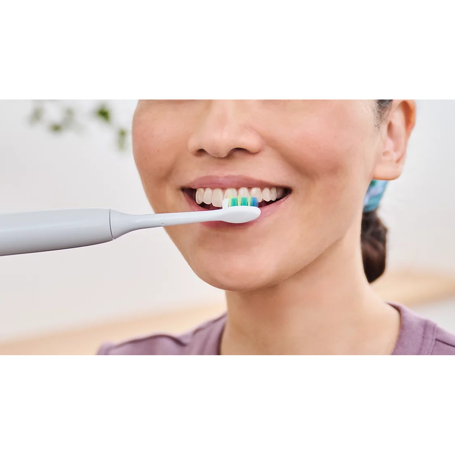 Una persona si lava i denti con uno spazzolino elettrico grigio in una stanza luminosa.