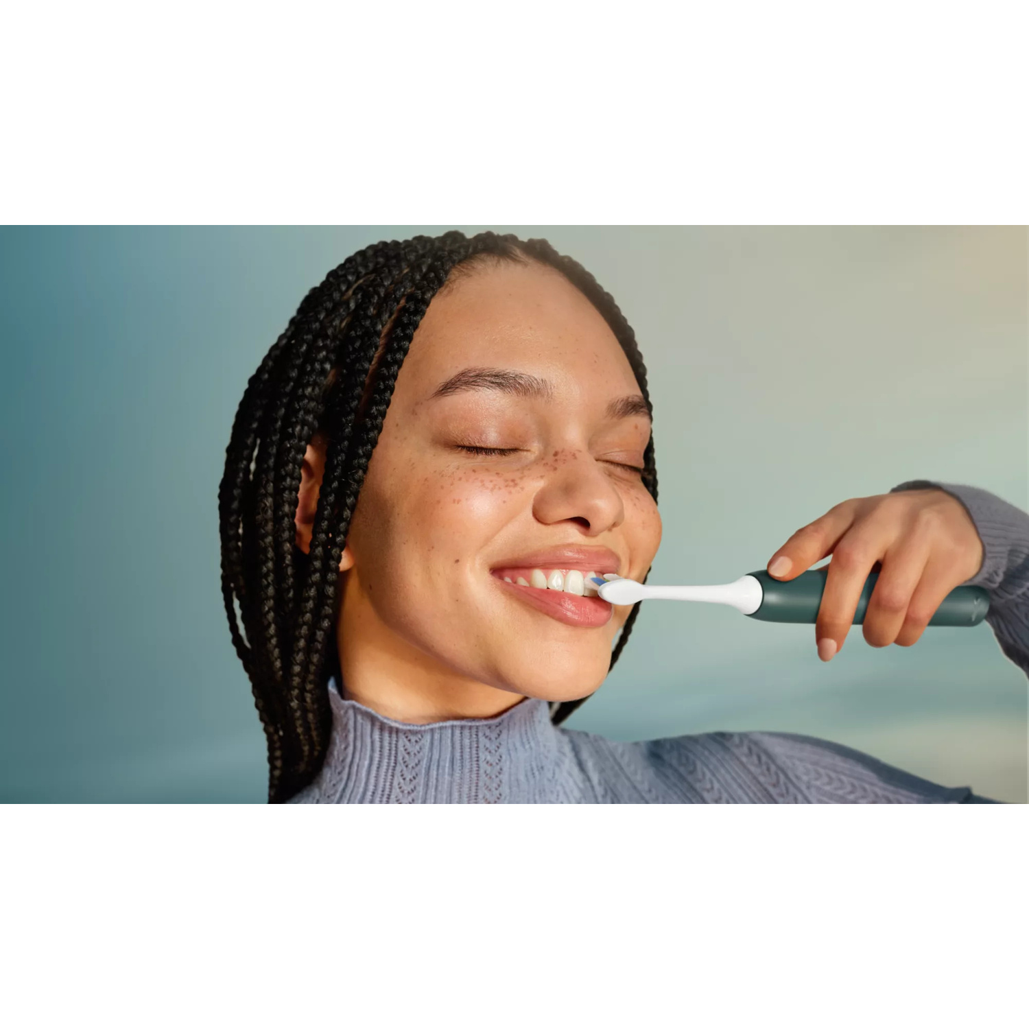 Una donna con trecce nere si lava i denti con uno spazzolino elettrico su sfondo blu.