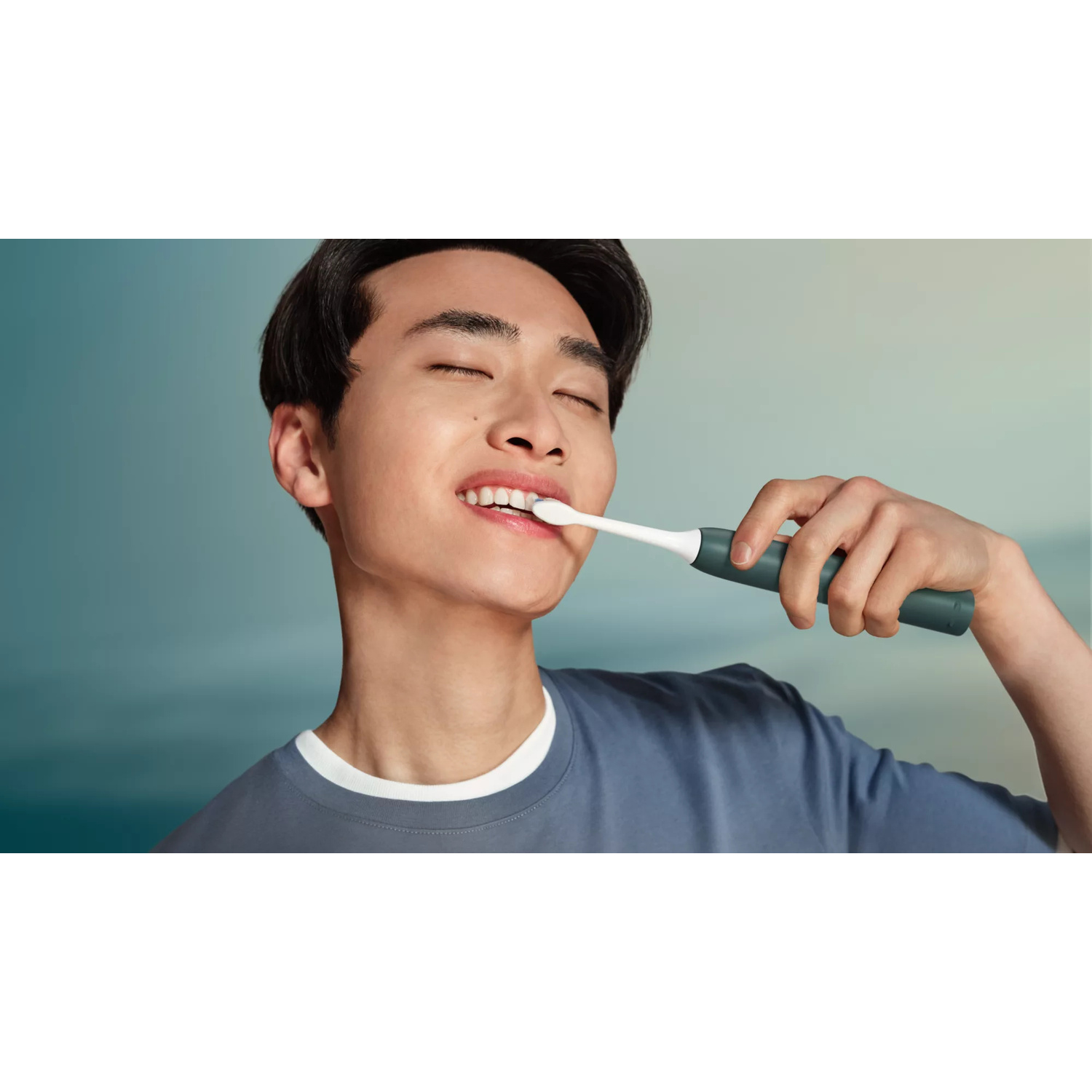 Un giovane uomo con gli occhi chiusi si lava i denti con uno spazzolino elettrico grigio scuro.