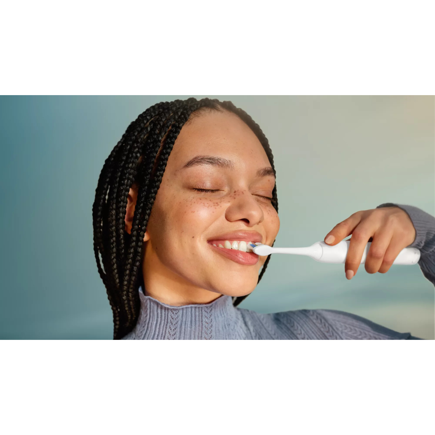 Una giovane donna con lunghe trecce si lava i denti usando uno spazzolino elettrico bianco.