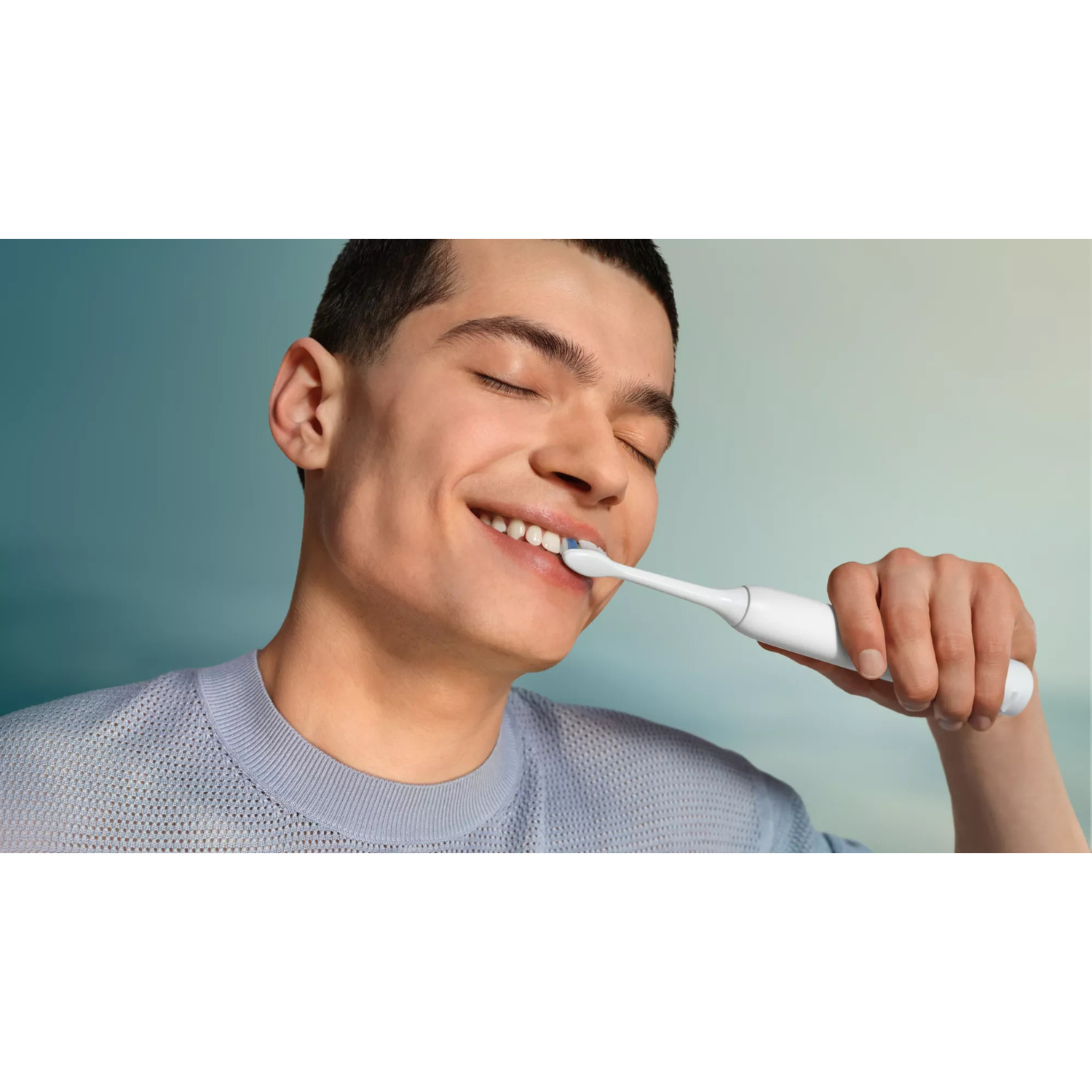 Un giovane uomo che indossa un maglione blu si lava i denti con uno spazzolino elettrico.