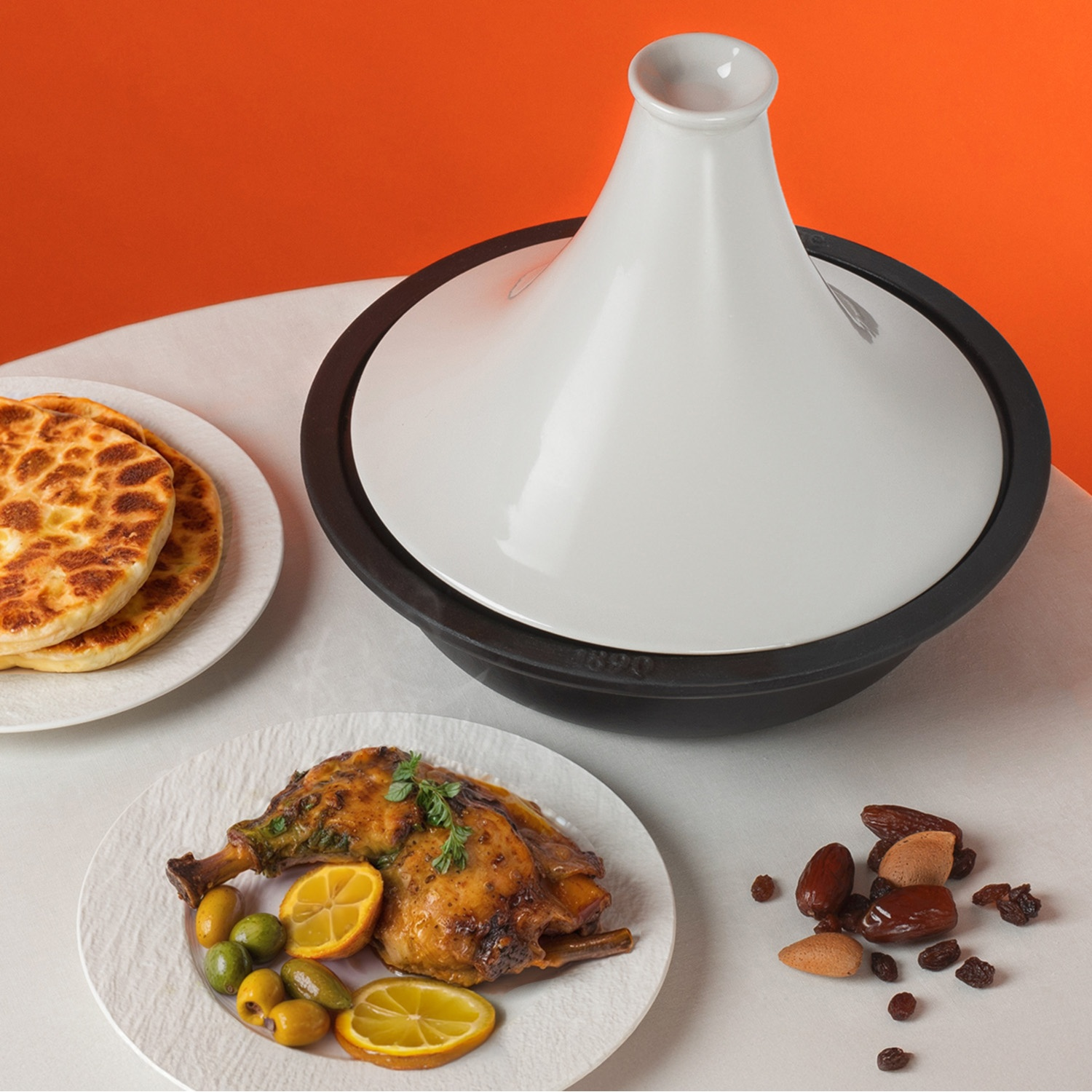 Una tajine bianca, coscia di pollo arrosto con olive, focaccia e noci su un tavolo con sfondo arancione.