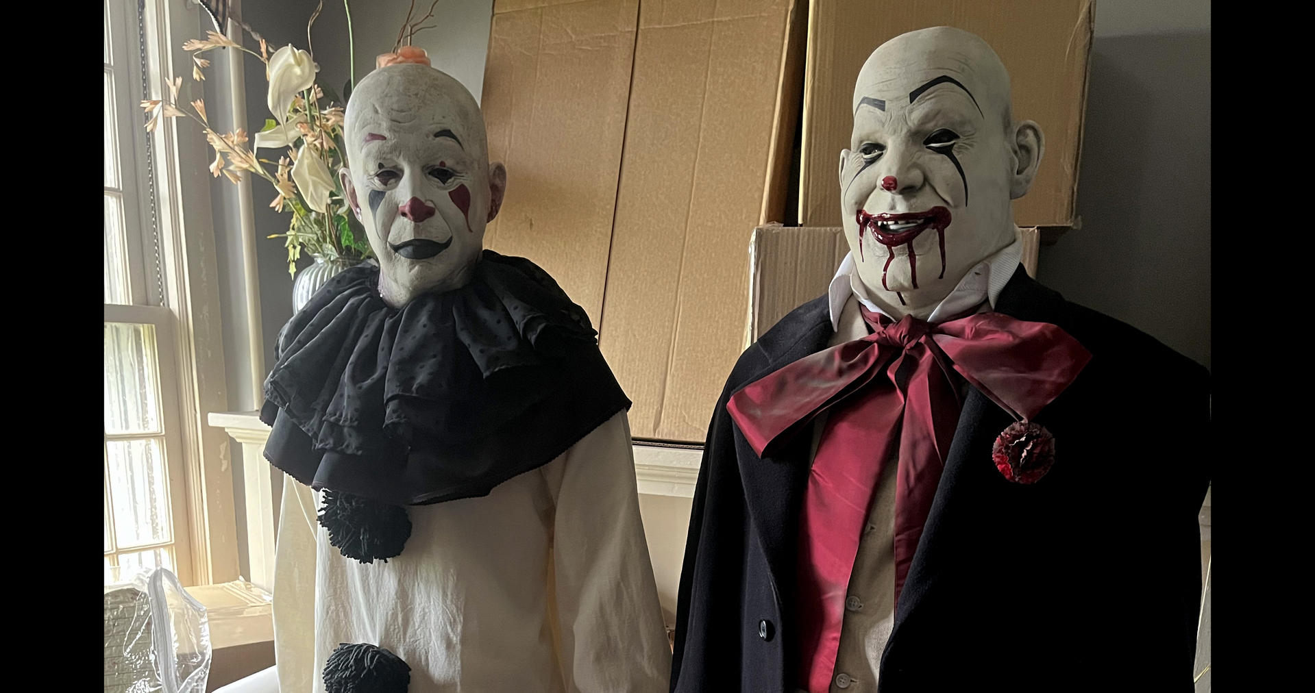 Zwei gruselige Horror-Clown-Puppen stehen vor Pappkartons in einem Innenraum.
