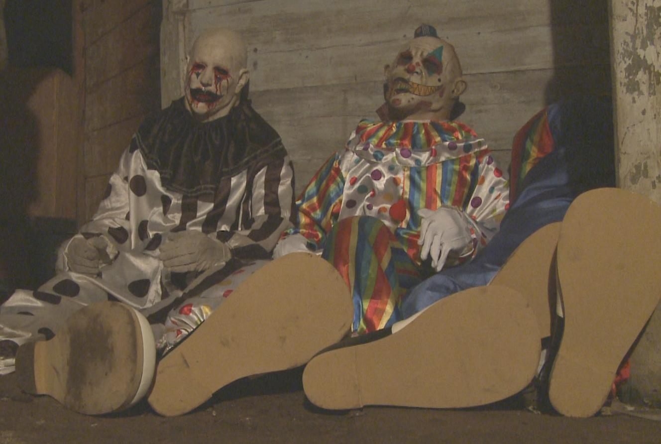 Drei gruselige Clown-Figuren sitzen mit großen übergroßen Schuhen in einer dunklen Kulisse an einer Wand.