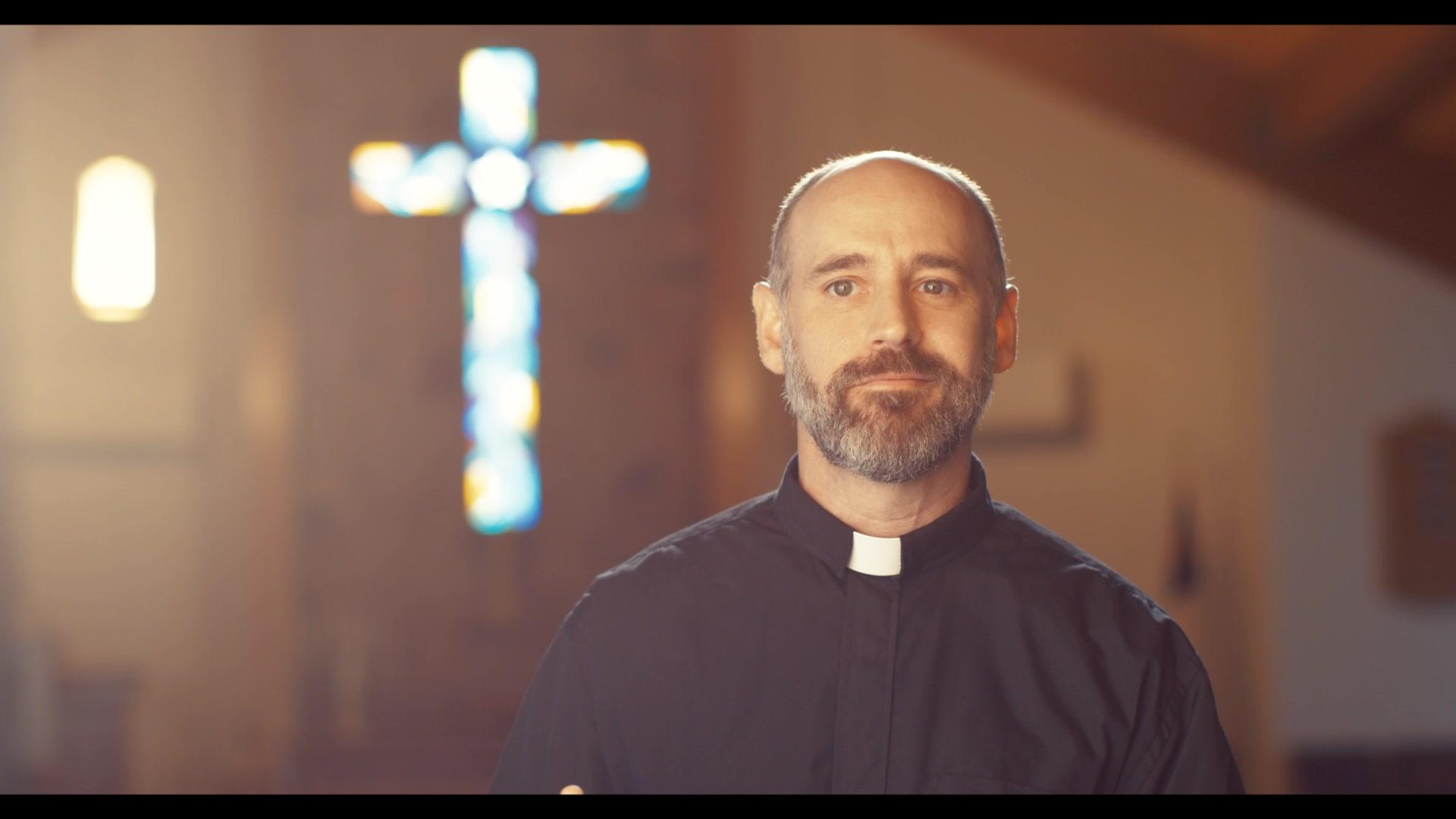 Ein Priester in schwarzem Hemd mit weißem Kragen steht in einer Kirche vor einem leuchtenden Kreuz.