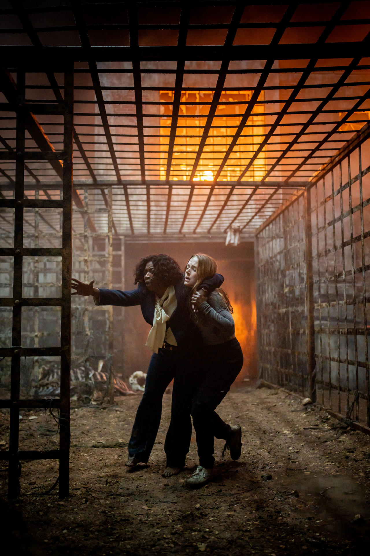 Zwei Frauen entkommen brennenden Metallkäfigen in einem dunklen Tunnel.