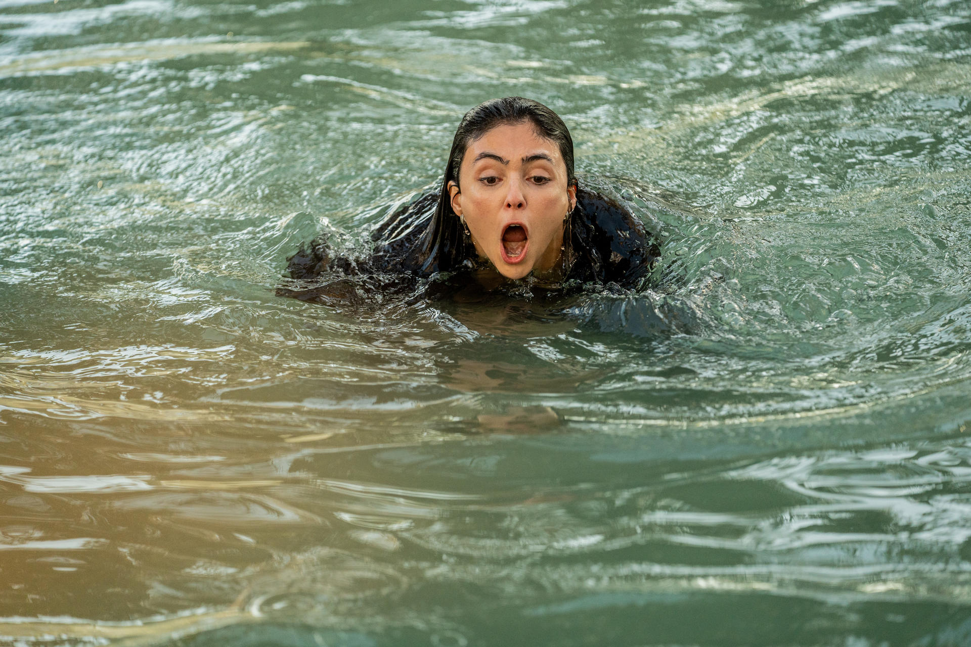 Eine Frau mit nassem dunklem Haar schwimmt im klaren Wasser und ruft mit weit geöffnetem Mund.