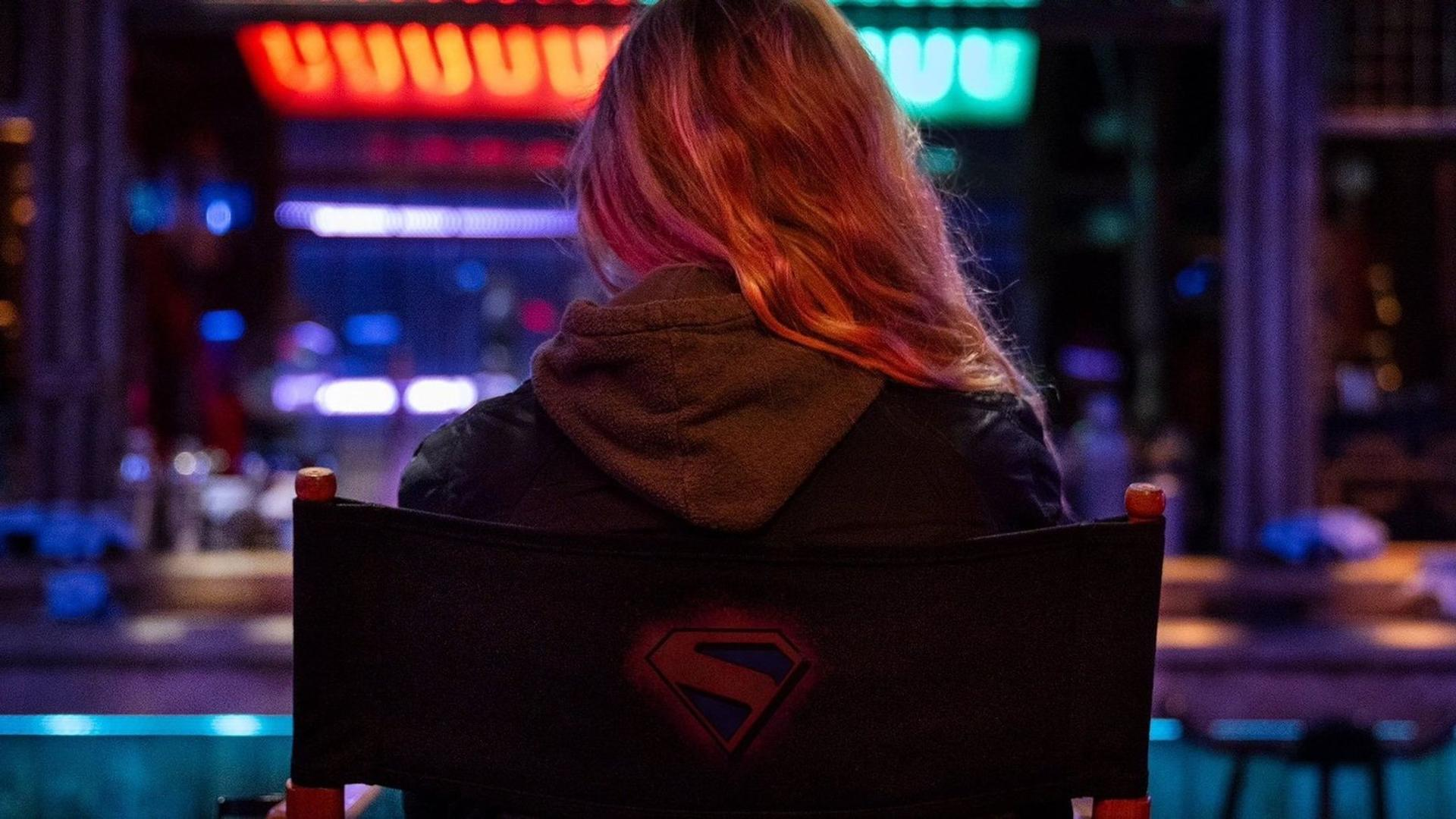 Frau mit blonden Haaren sitzt von hinten in einem Stuhl mit Superman-Logo vor neonbeleuchtetem Hintergrund.
