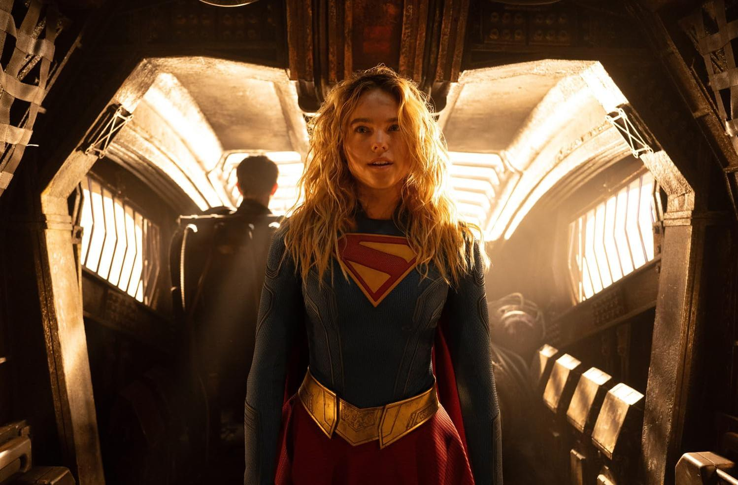 Supergirl mit blondem Haar und blau-rotem Anzug steht in einem Raumschiff, im Hintergrund eine Person.