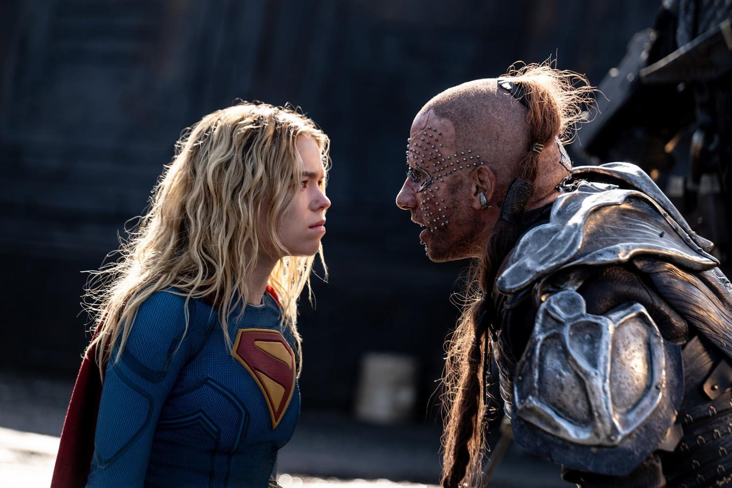 Supergirl mit blonden Haaren blickt einem bärtigen Krieger mit Metallrüstung und Gesichtspiercings intensiv in die Augen.