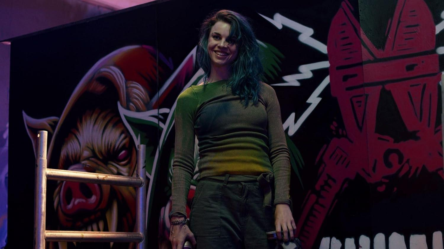 Eine junge Frau mit blauen Haaren steht lächelnd vor einer bunten Graffiti-Wand.