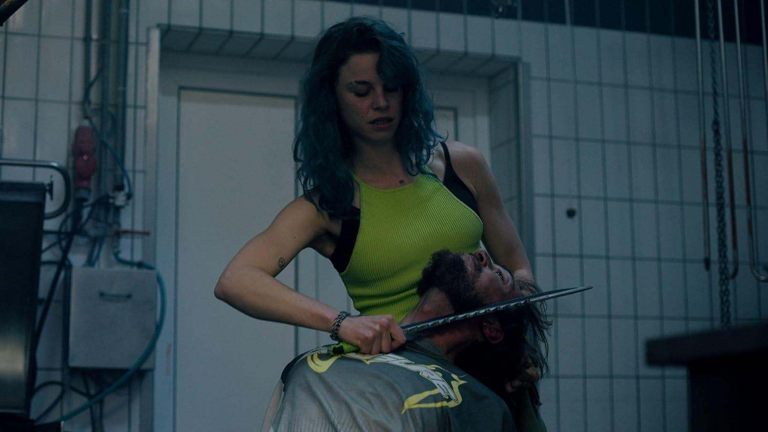 Eine blauhaarige Frau hält in einem gefliesten Raum ein Messer an den Hals eines Mannes.
