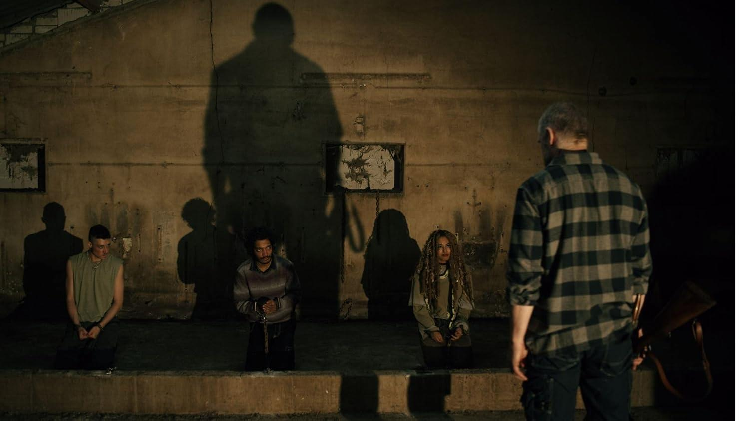 Ein bewaffneter Mann steht vor drei gefesselten Gefangenen vor einer Wand mit Schatten.